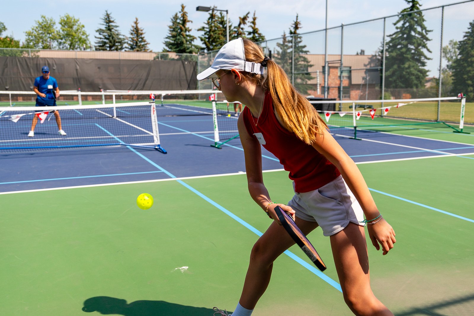A young woman in a red tank top and white shorts playing pickleball on an outdoor court, preparing to hit a yellow ball with a paddle. A man is in the background near the net. The court is fenced, and there are trees and buildings around.