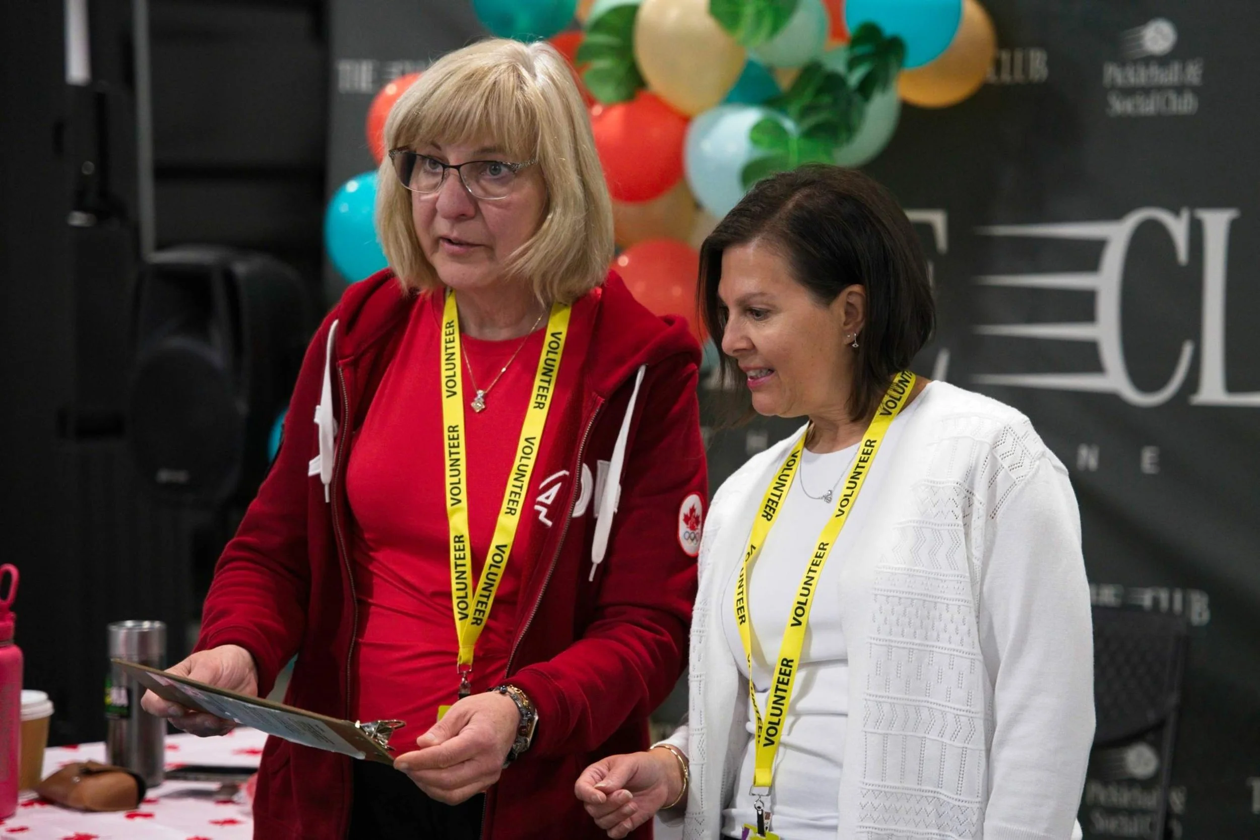 Two women wearing yellow 'Volunteer' lanyards, standing indoors at a table with colorful balloons in the background, looking at documents.