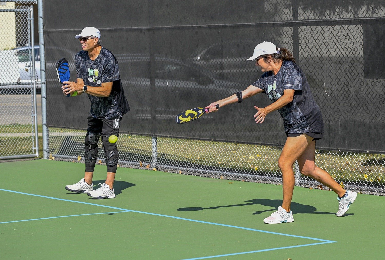 Two women playing pickleball on a green outdoor court with a black windscreen and chain-link fence in the background. One woman is holding a paddle and appears to be preparing to hit a yellow ball, while the other woman watches.