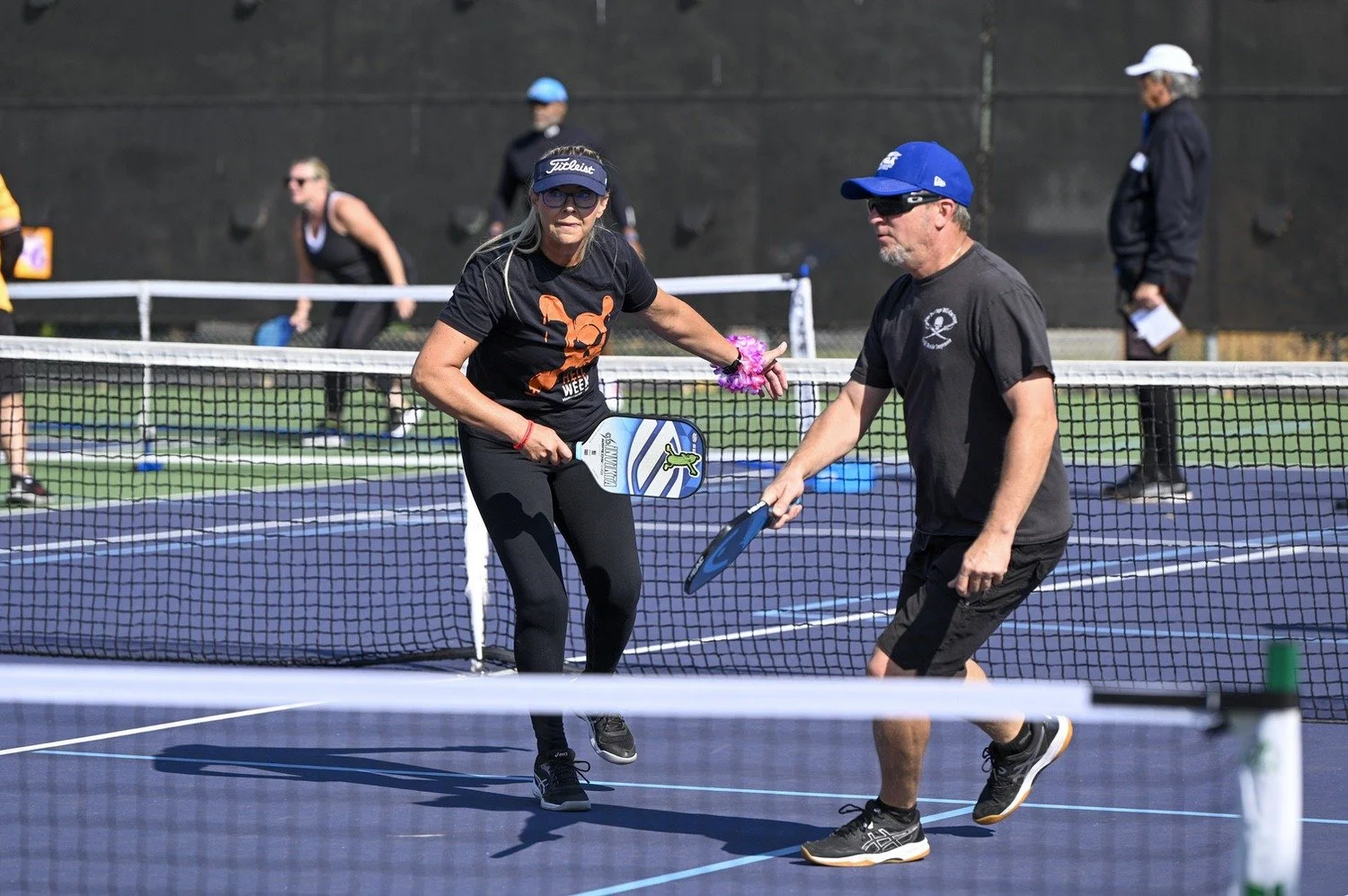 People playing pickleball on an outdoor court. Two players are in the foreground, holding paddles and engaging in a game. Additional players and referees are visible in the background.