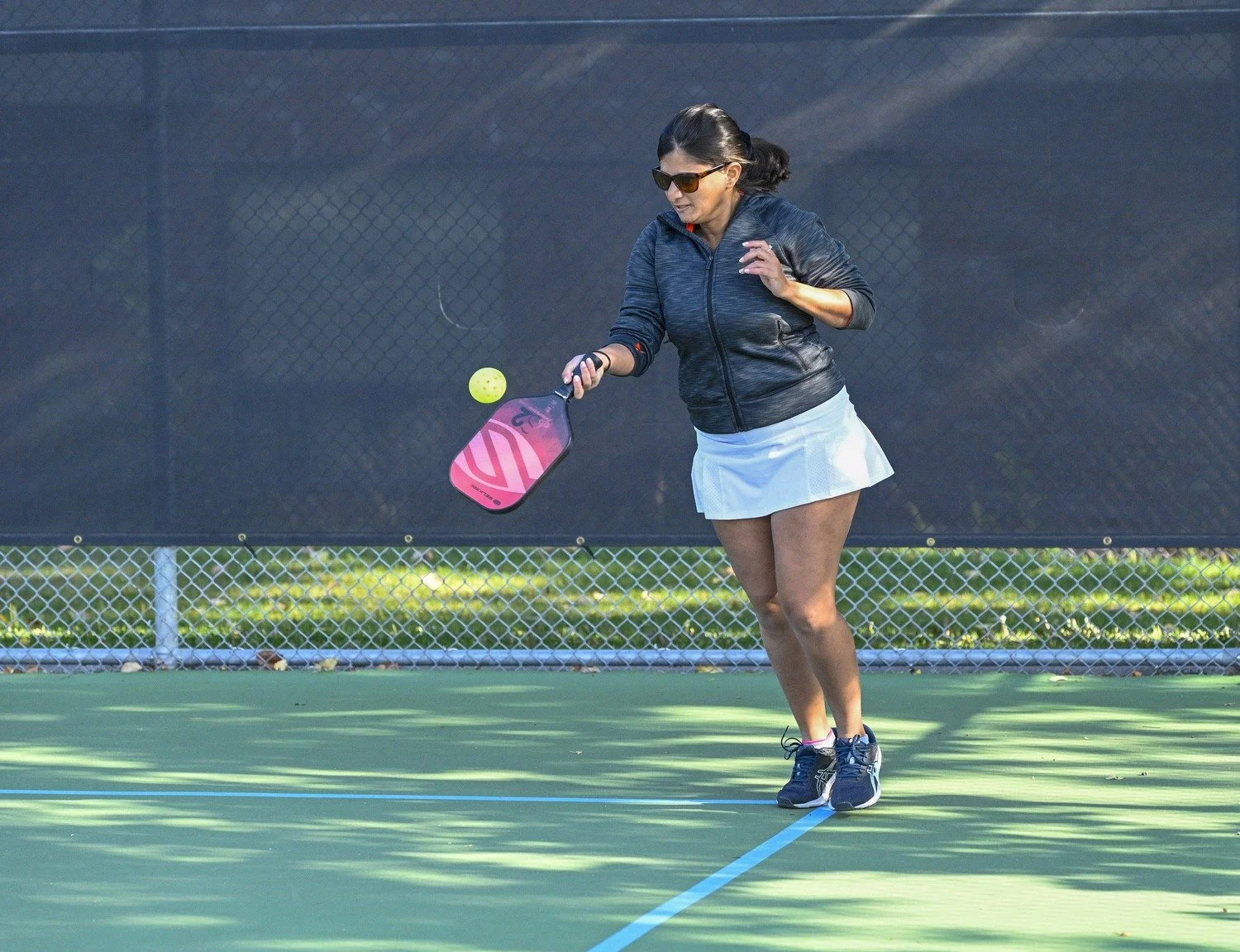 A woman playing pickleball on an outdoor court, wearing sunglasses, a black jacket, and a white skirt, hitting a yellow pickleball with a pink paddle.