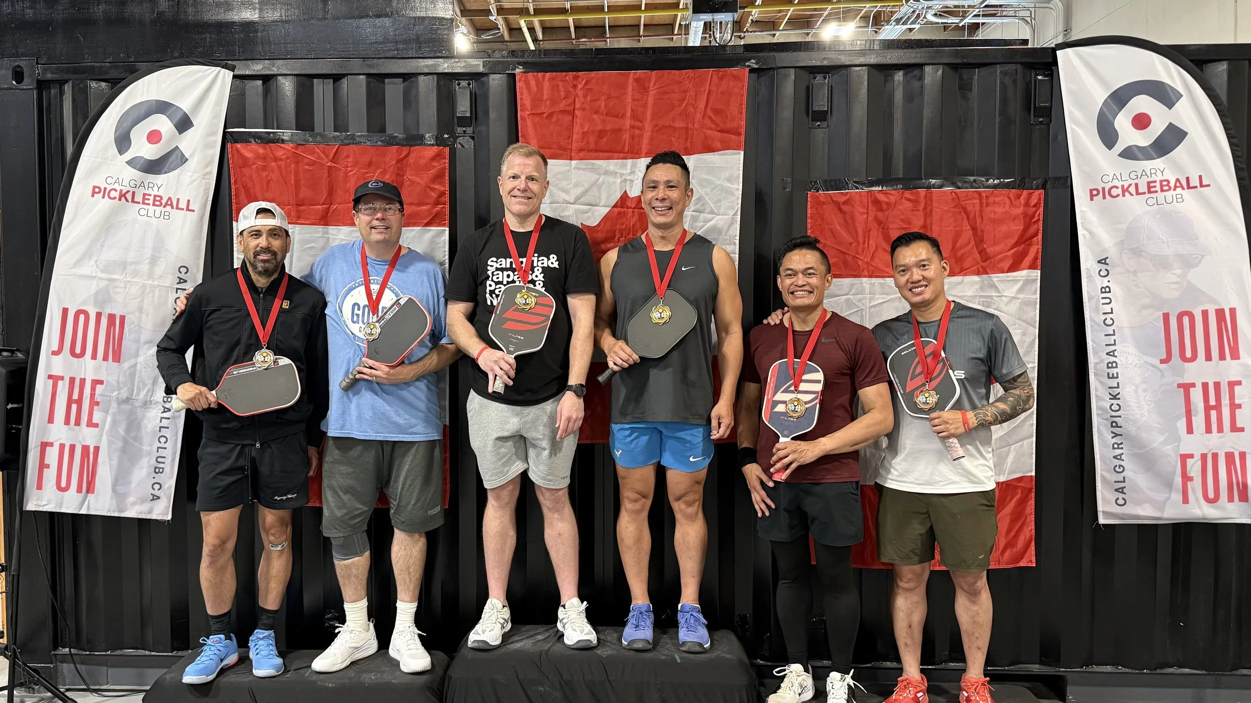A group of six men standing together on a platform, holding paddle-shaped awards, with medals around their necks, at a ping pong event hosted by Calgary Pickleball Club. They are smiling, and behind them are flags with the Calgary Pickleball Club log