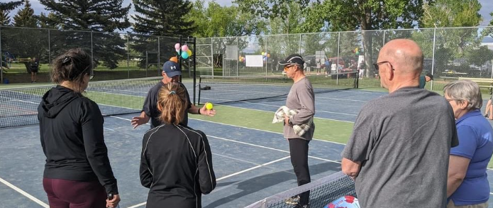 Group of people gathered on a tennis court, with some holding tennis balls and a woman in the center demonstrating or instructing.
