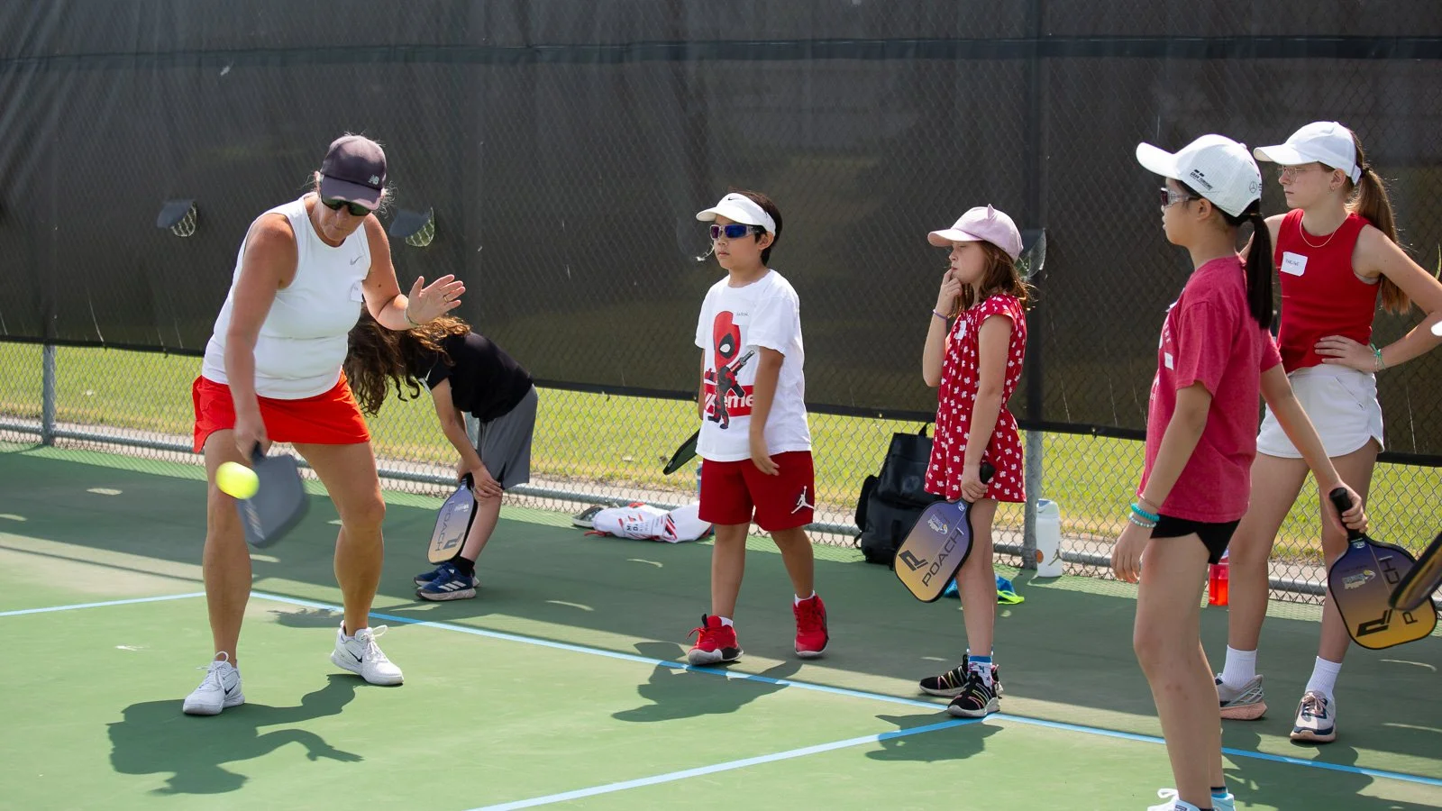 Instructor demonstrating tennis technique to young children on a tennis court, with children holding tennis paddles and watching attentively.