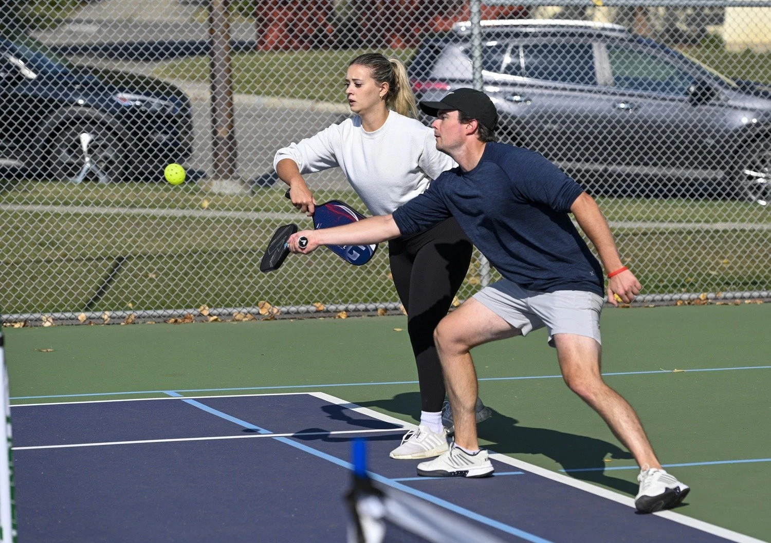 A man and a woman playing pickleball on an outdoor court enclosed by a chain-link fence, with parked cars in the background. The man is hitting a yellow pickleball with a paddle, and the woman is preparing to hit it back.