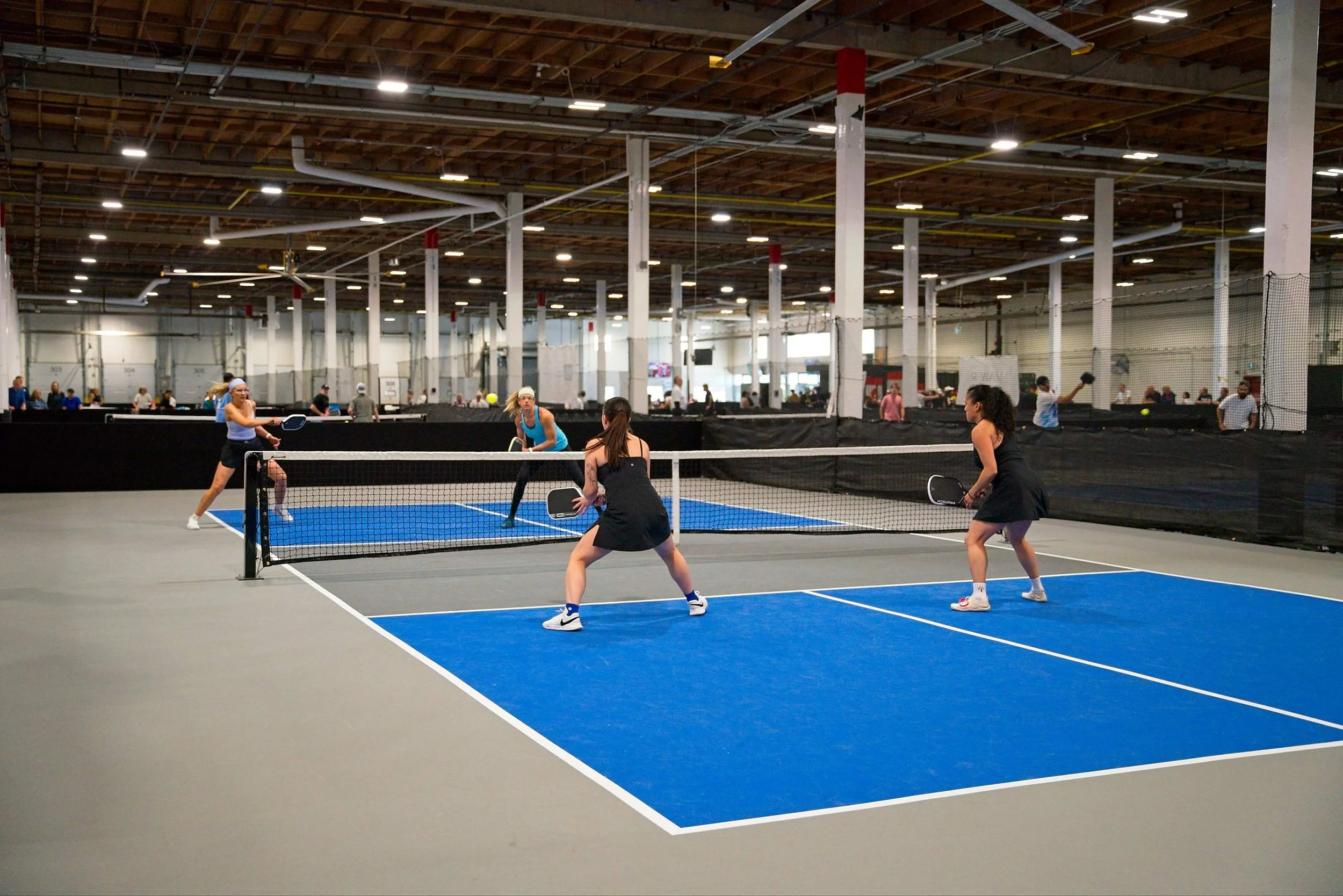 People playing pickleball on indoor court with blue surface, basketball net, and net dividing court