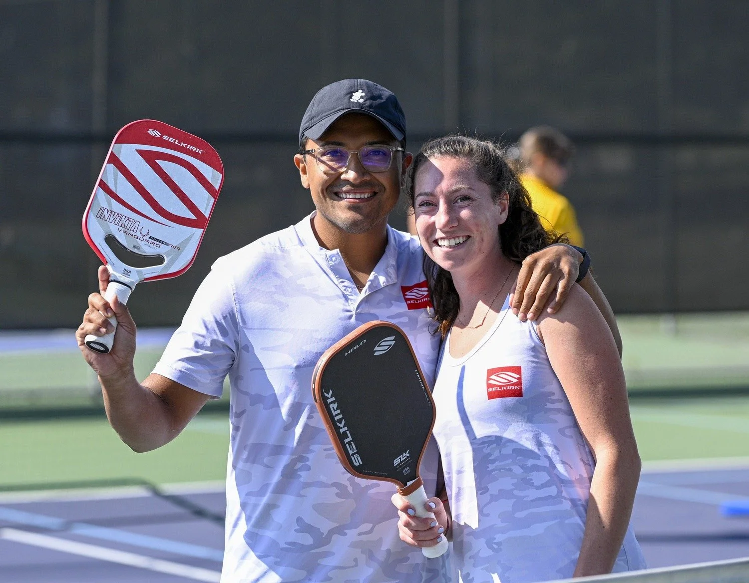 Two people smiling and holding pickleball paddles on a paddle court, with one person having an arm around the other's shoulders.