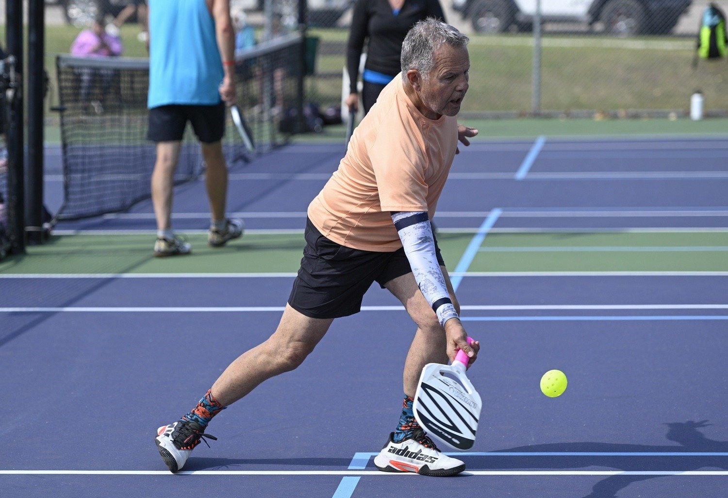 Older man playing pickleball on a blue court, reaching to hit a yellow ball with a paddle, wearing a coral shirt, black shorts, and multicolored socks.
