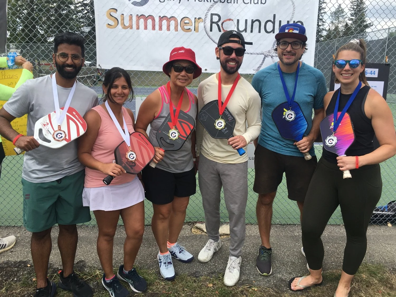 Group of six people smiling with medals and paddle racquets, standing outdoors in front of a chain-link fence and a banner that reads 'Summer Roundup'.