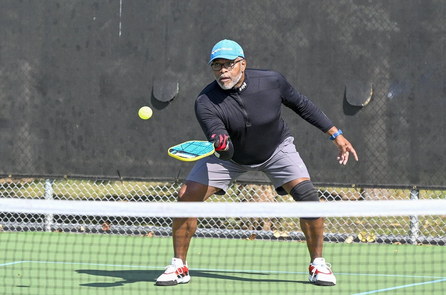 An older man playing pickleball on an outdoor court, wearing a blue cap, glasses, black long sleeve shirt, gray shorts, and sneakers, holding a pickleball paddle and reaching for a yellow ball.