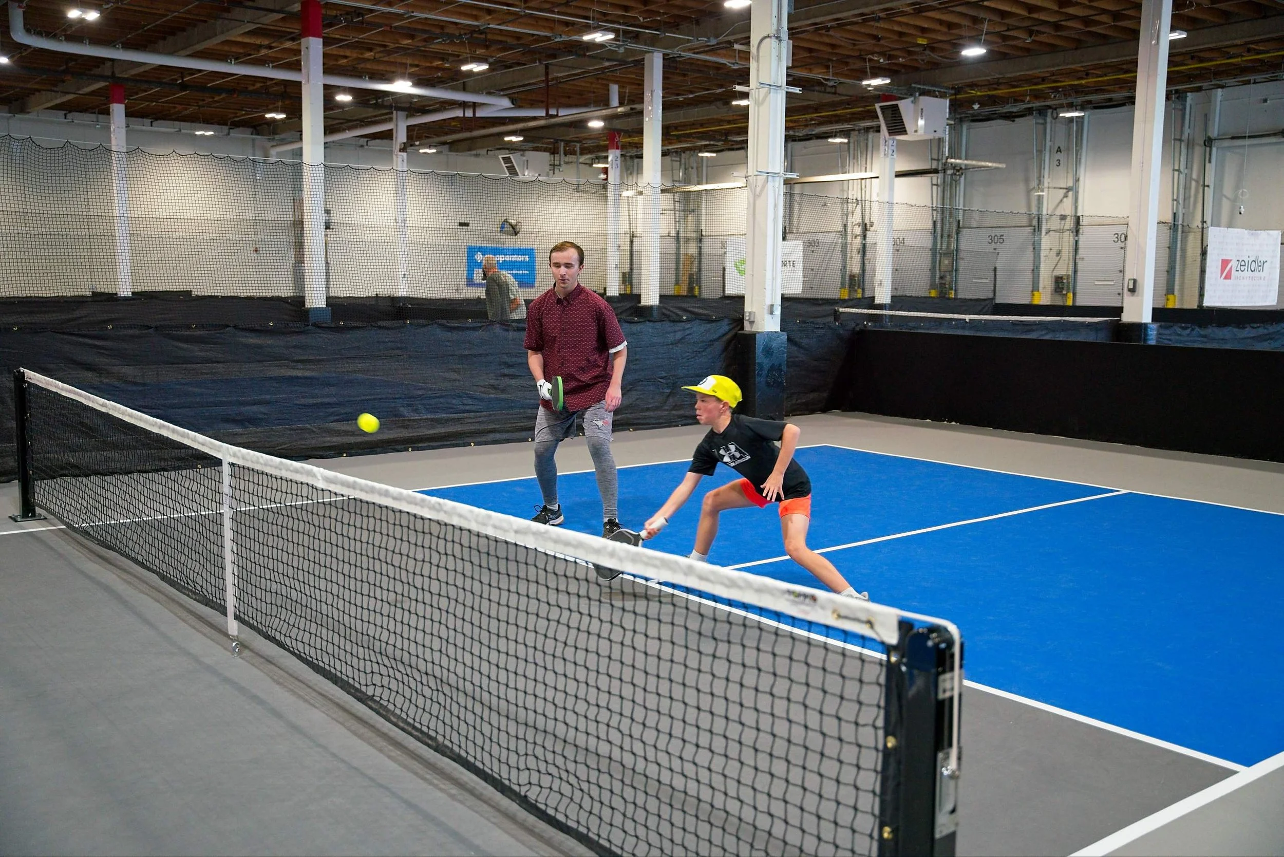 Two boys playing tennis on an indoor court, with a man watching in the background. The court has a blue surface and a black net, with a warehouse-style ceiling and overhead lighting.