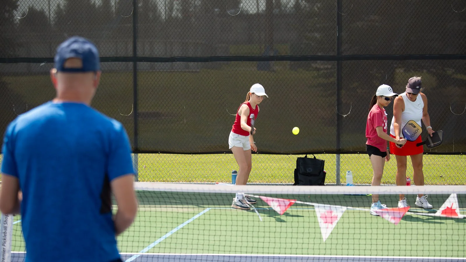 People playing pickleball on an outdoor court. A man in a blue shirt and cap is in the foreground with his back to the camera, while three women are on the court, with one in a white cap preparing to hit the ball, and another woman in a black cap and