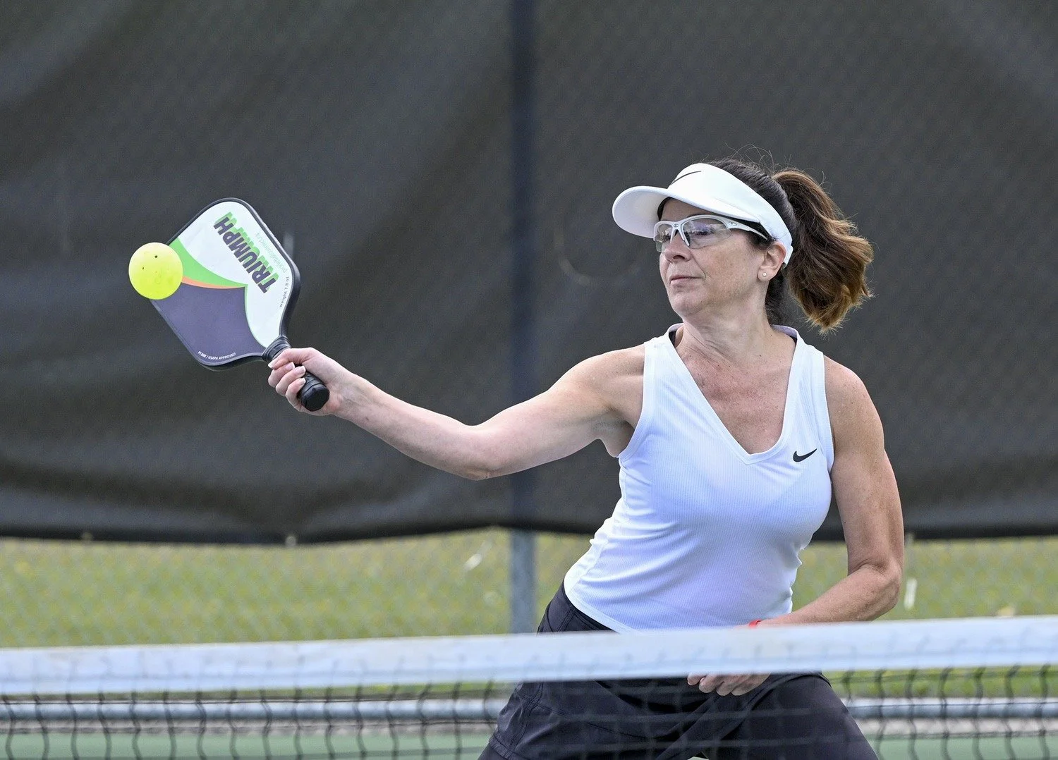 A woman playing pickleball on an outdoor court, wearing sportswear, goggles, and a visor, hitting a yellow pickleball with a paddle.