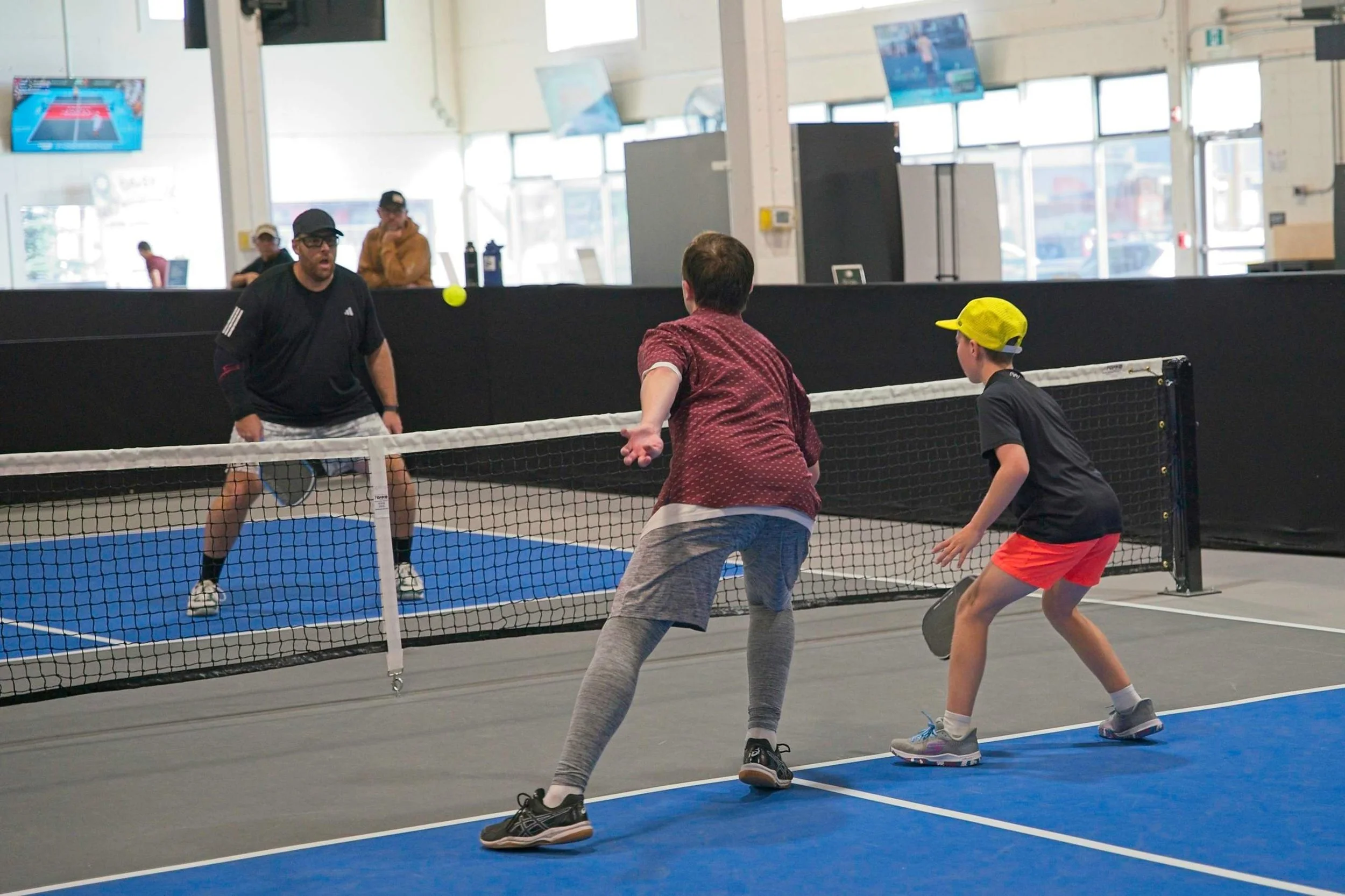 Three people playing pickleball indoors, with a black net and blue court, watching a yellow tennis ball.