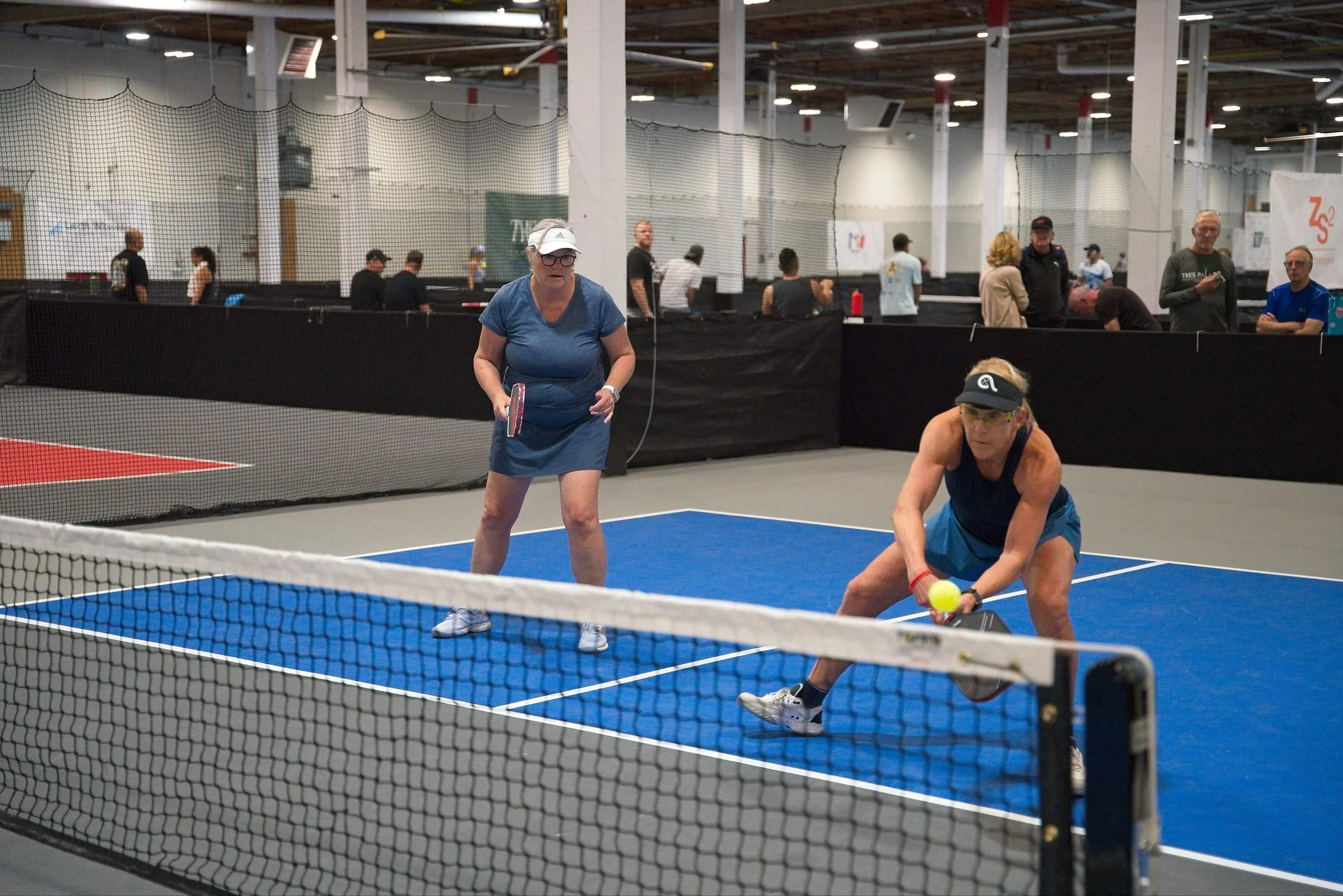 Two older women playing pickleball on an indoor court, one preparing to hit a yellow ball while the other stands nearby holding a paddle, with spectators and other players in the background.