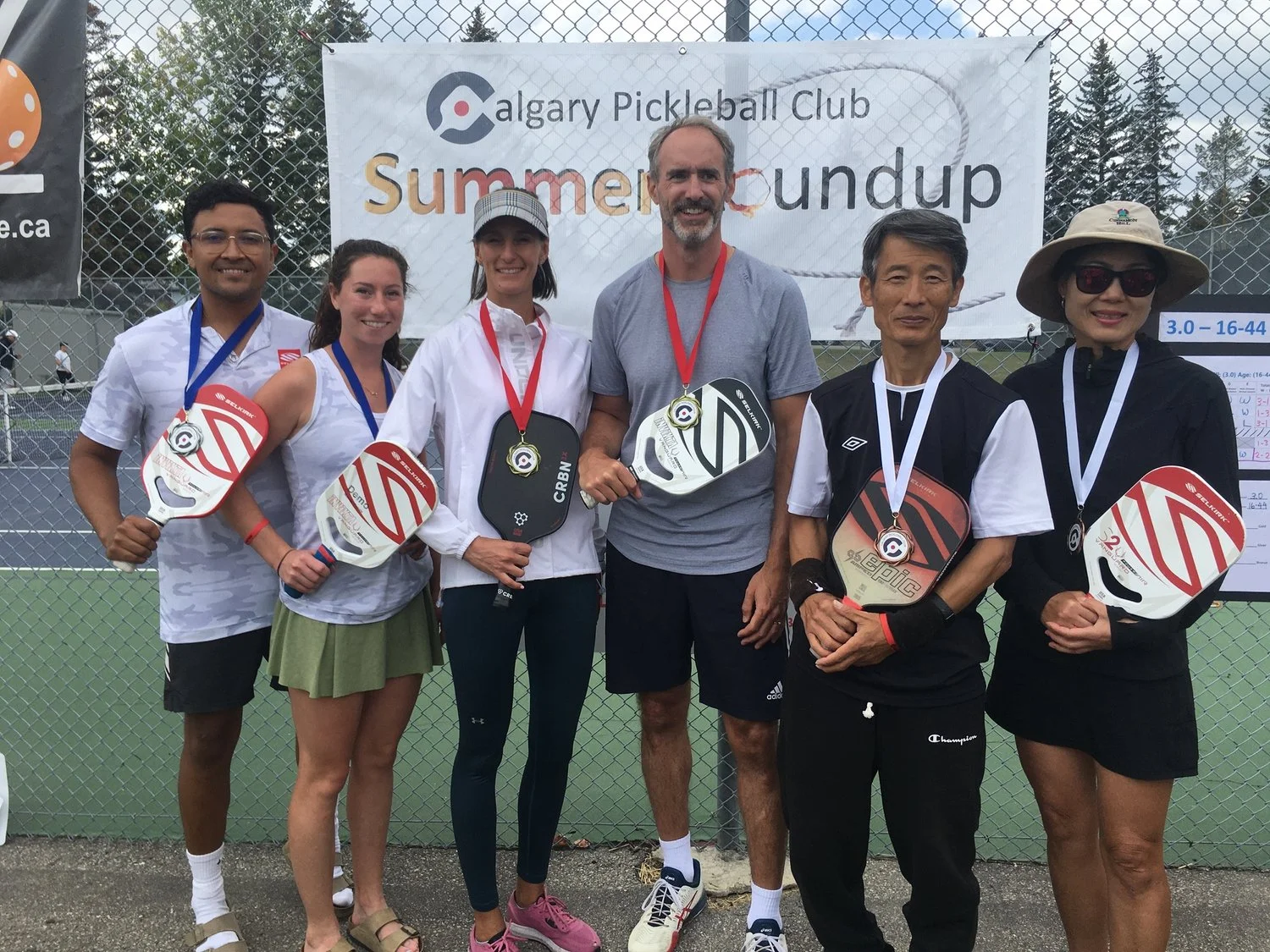 Group of seven people standing in front of a net on a tennis court, holding pickleball paddles and wearing medals, during a summer pickleball tournament organized by Calgary Pickleball Club.