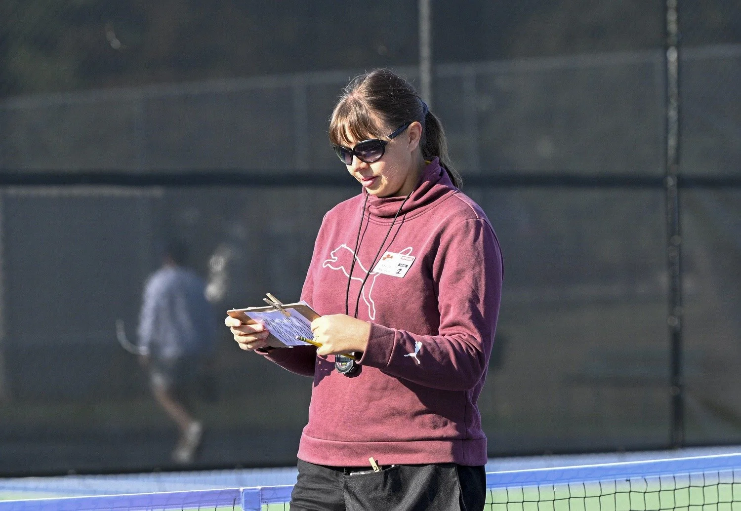 A woman with sunglasses and a maroon Puma hoodie writing on a clipboard at a tennis court.