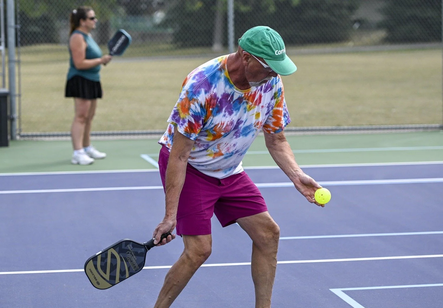 An elderly man in colorful tie-dye shirt and burgundy shorts holding a pickleball paddle in one hand and a yellow pickleball in the other on a pickleball court. A woman in a teal tank top and black skirt stands in the background.