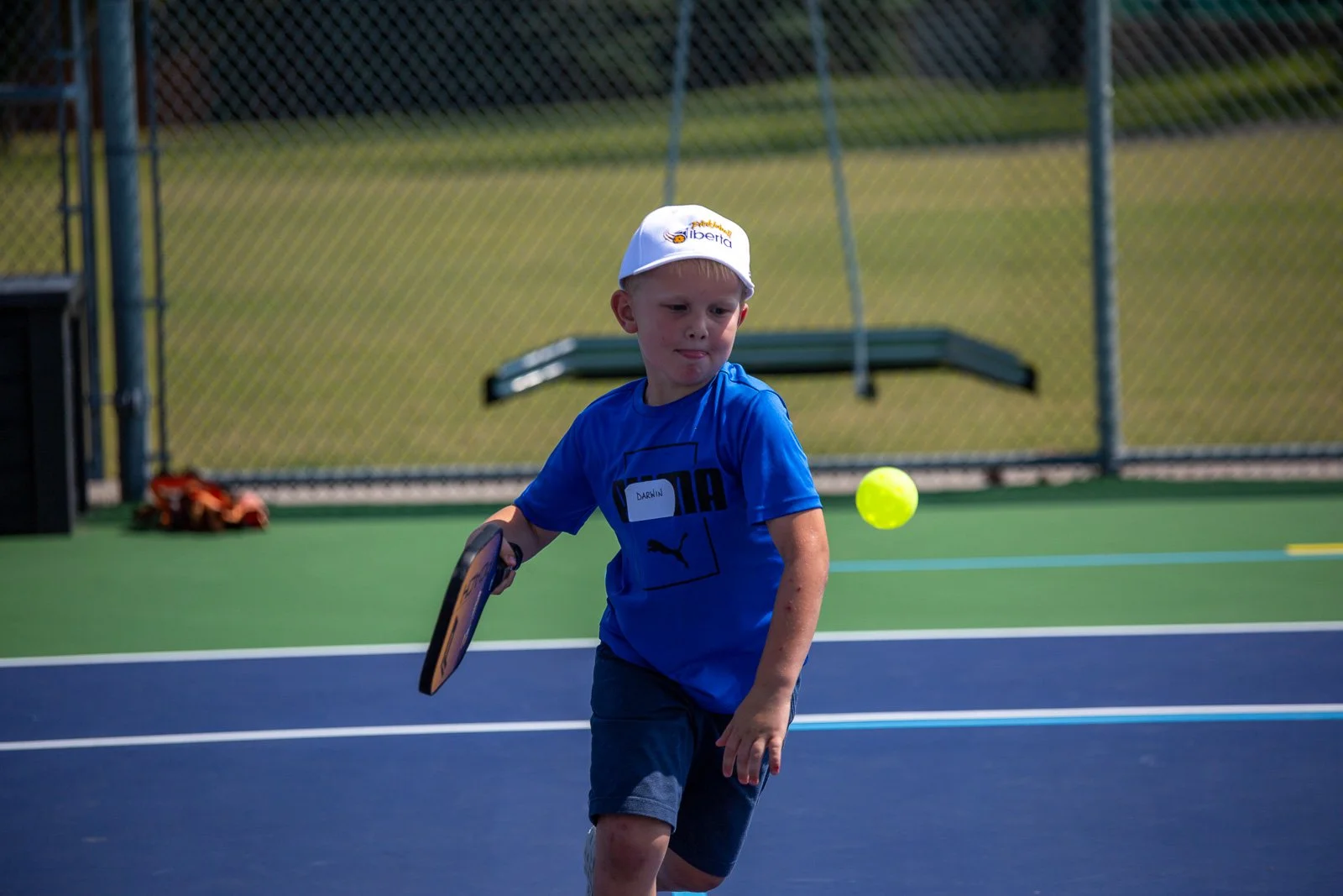 A young boy on a tennis court playing with a tennis ball and a racket, wearing a white cap and a blue shirt.