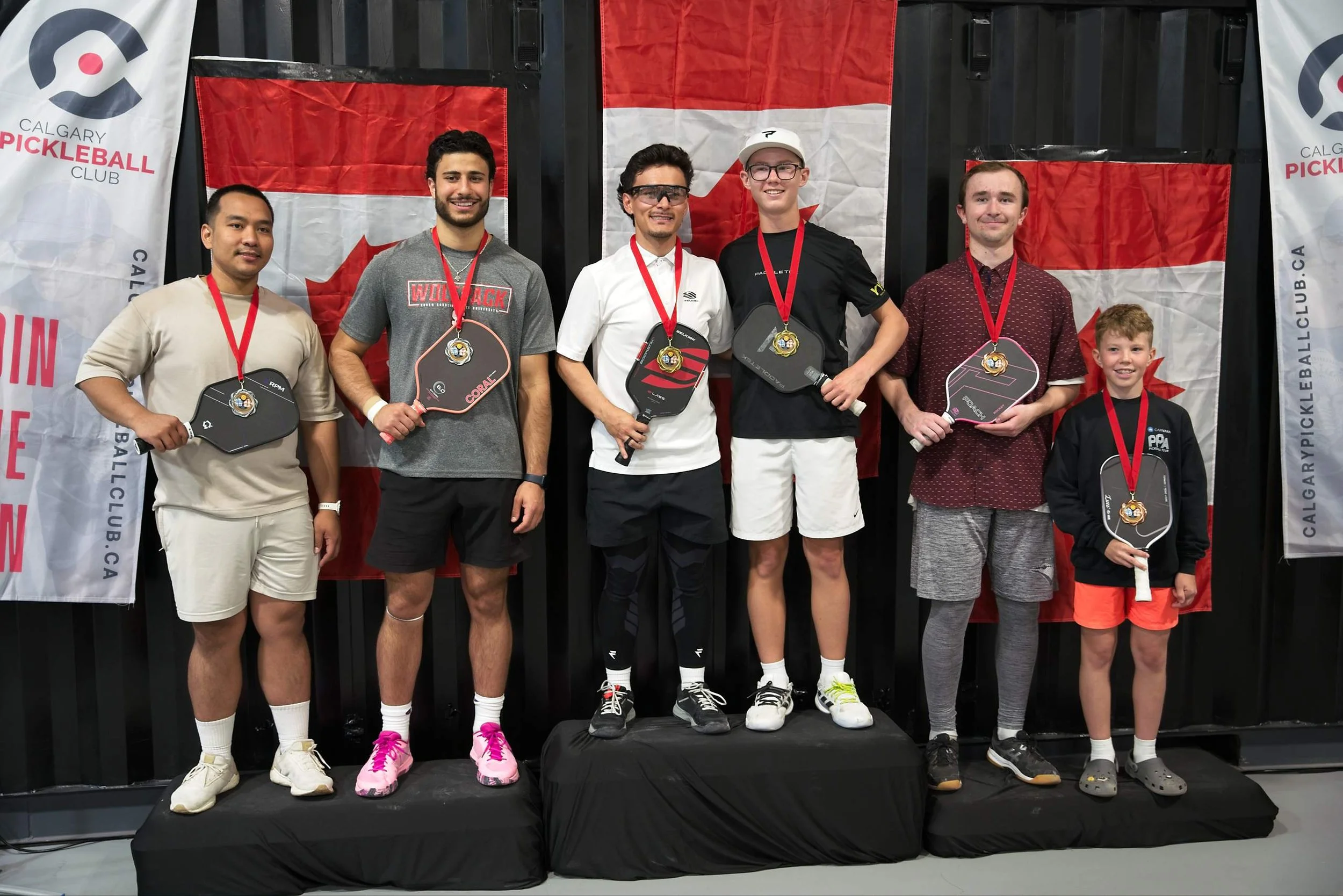 Group of five men and one boy standing on a podium after a pickleball competition, holding paddles and medals. Two Canadian flags and banner for Calgary Pickleball Club in the background.