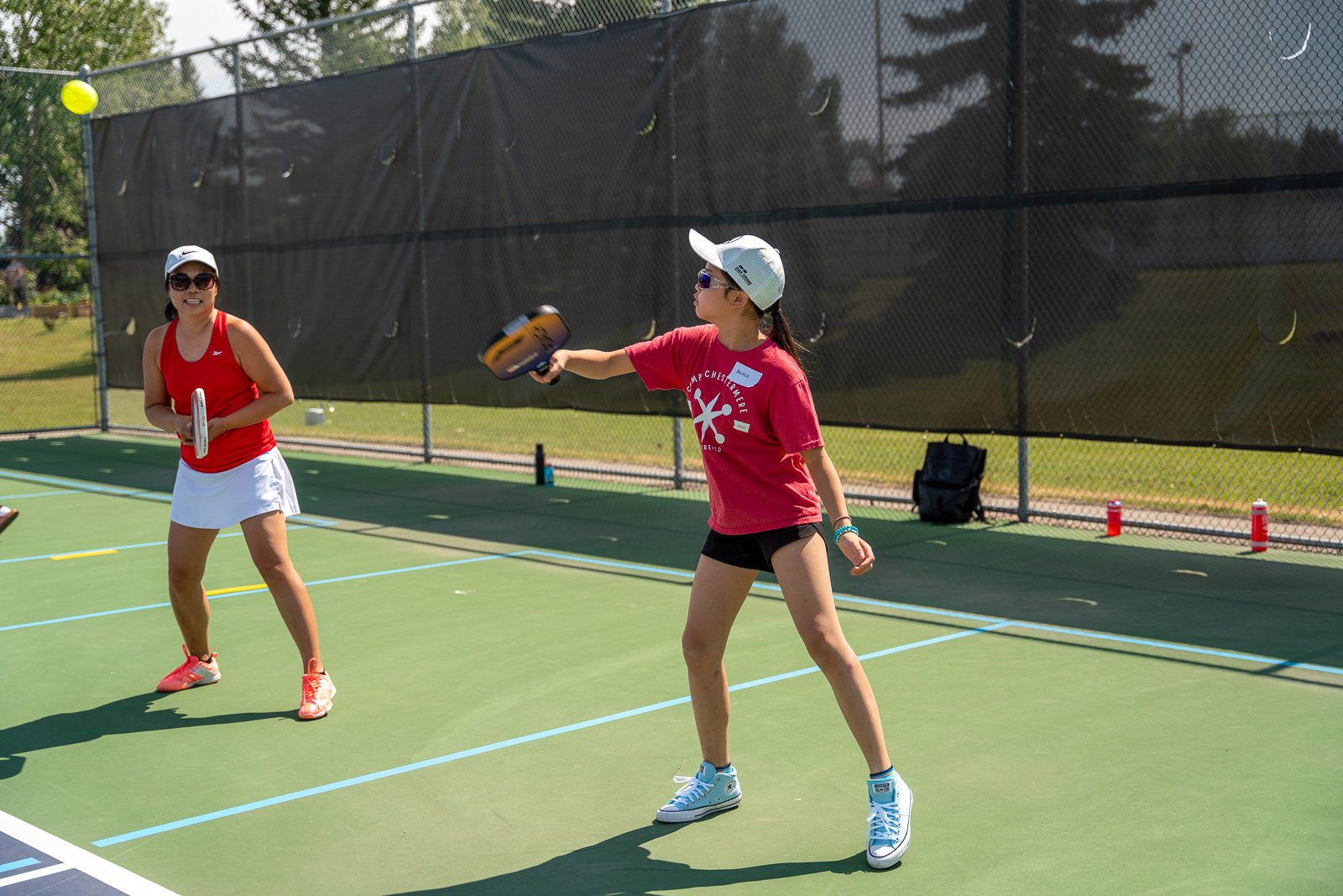 Two women playing pickleball on an outdoor court. One woman is preparing to hit the ball, while the other watches. They are wearing sporty clothes, hats, and sunglasses, with tennis shoes. The court is surrounded by a chainlink fence, and there are t