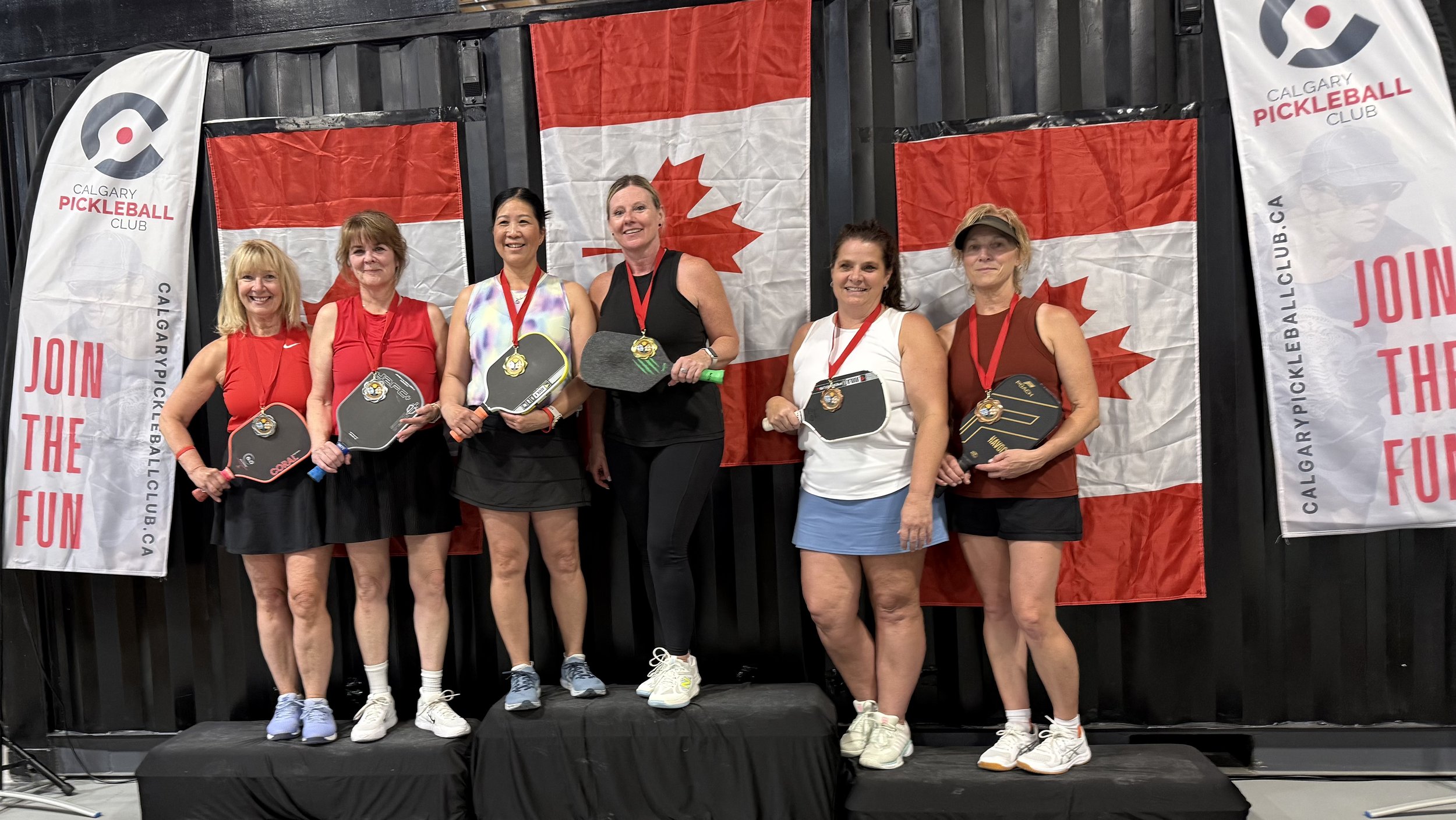 Six women standing on a stage, holding ping pong paddles and medals, with Canadian flags and banners for the Calgary Pickleball Club in the background.