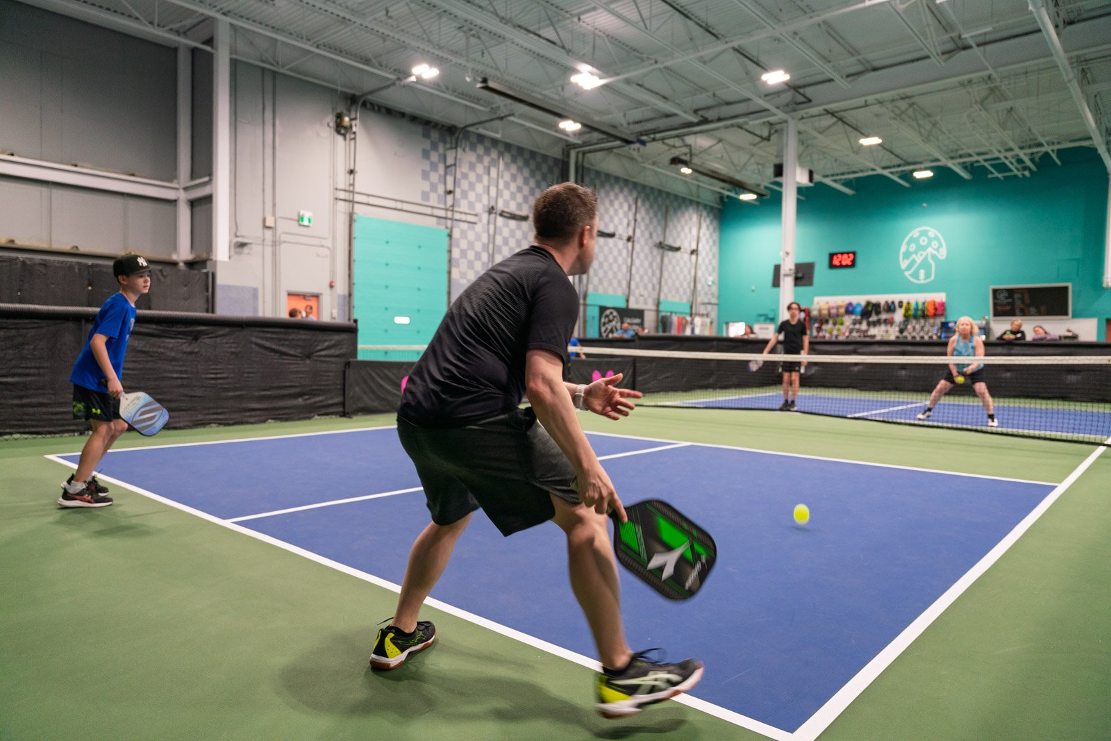 Group of people playing pickleball inside an indoor sports facility, with players ready to hit a yellow pickleball over a net.