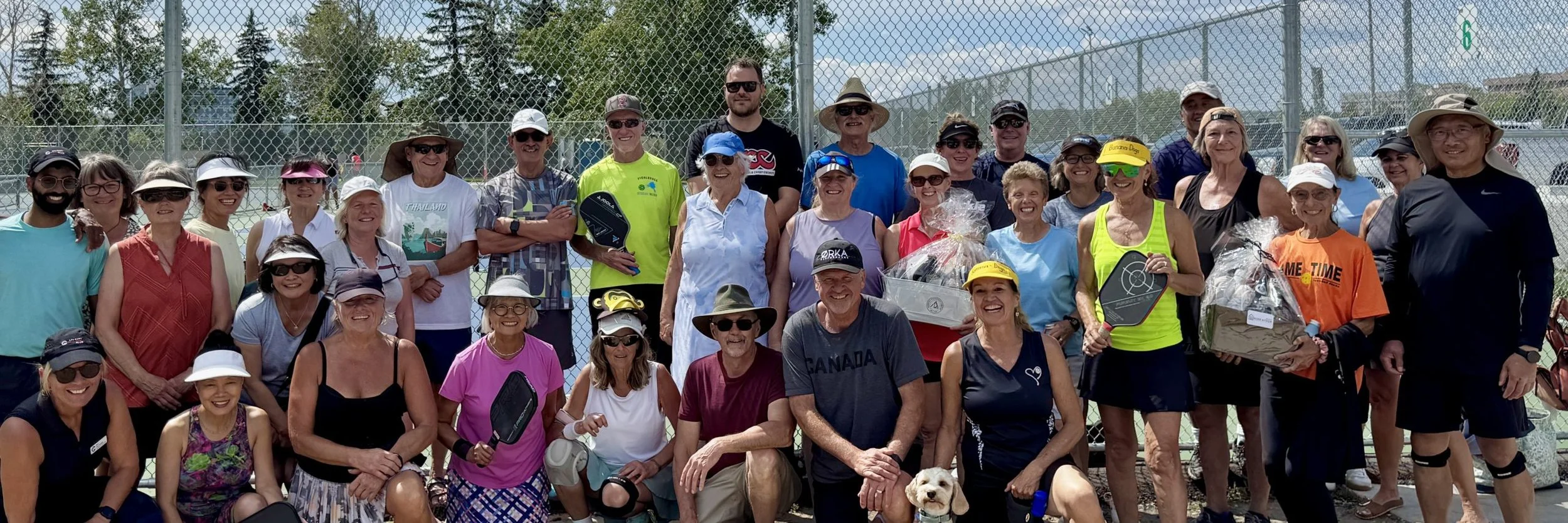Group of smiling people, mostly women, standing behind a tennis court fence, some holding paddles, in a sunny outdoor setting.