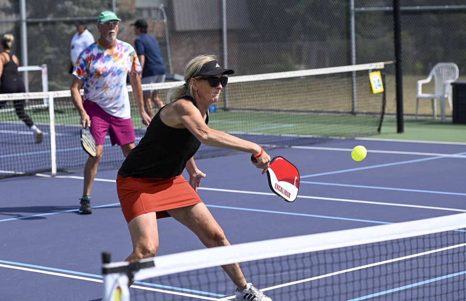 A woman playing pickleball on an outdoor court, hitting a yellow ball with a paddle, with a man in colorful clothing behind her and other people in the background.