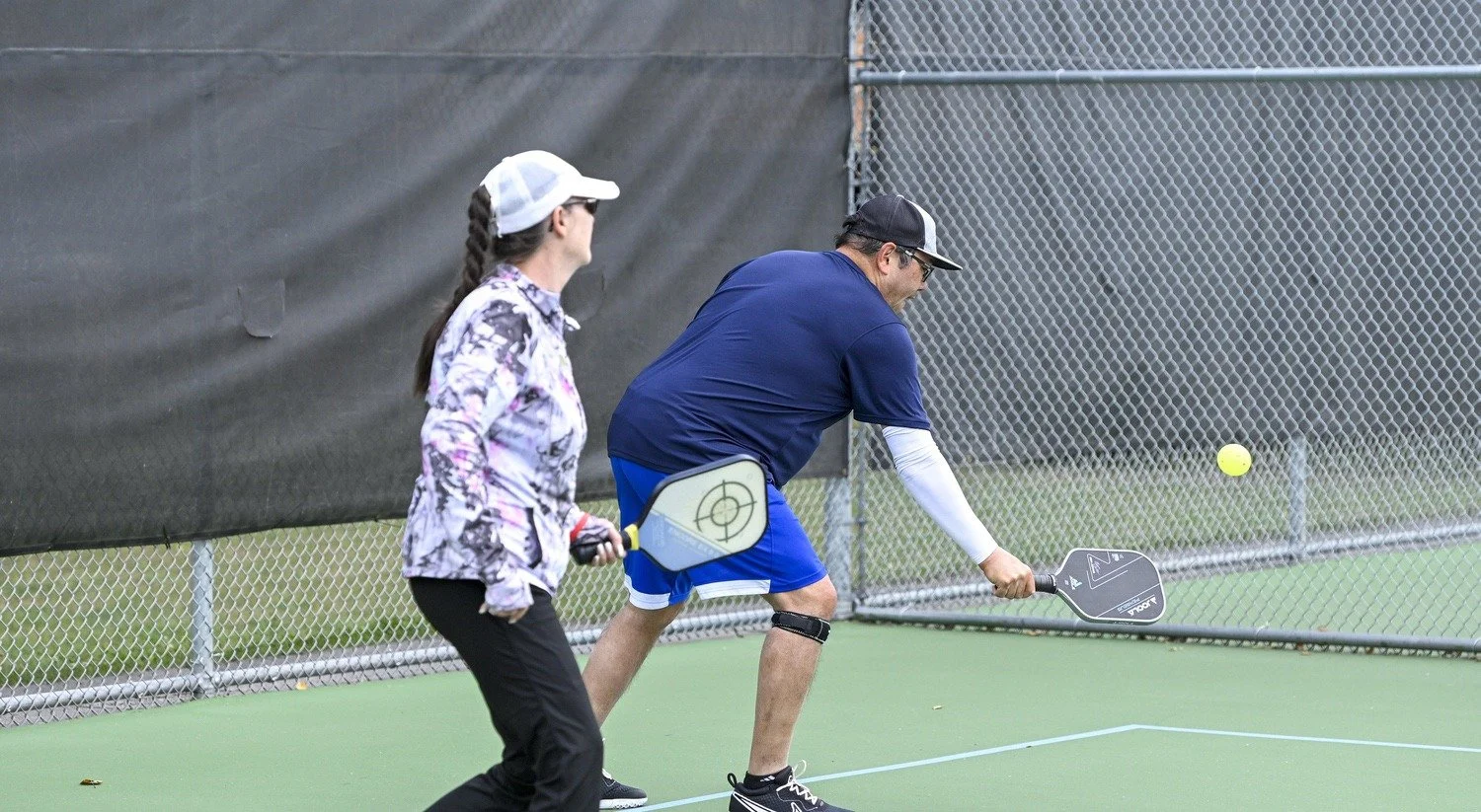Two people playing pickleball on a court. One person is about to hit a yellow ball with a paddle, while the other watches.
