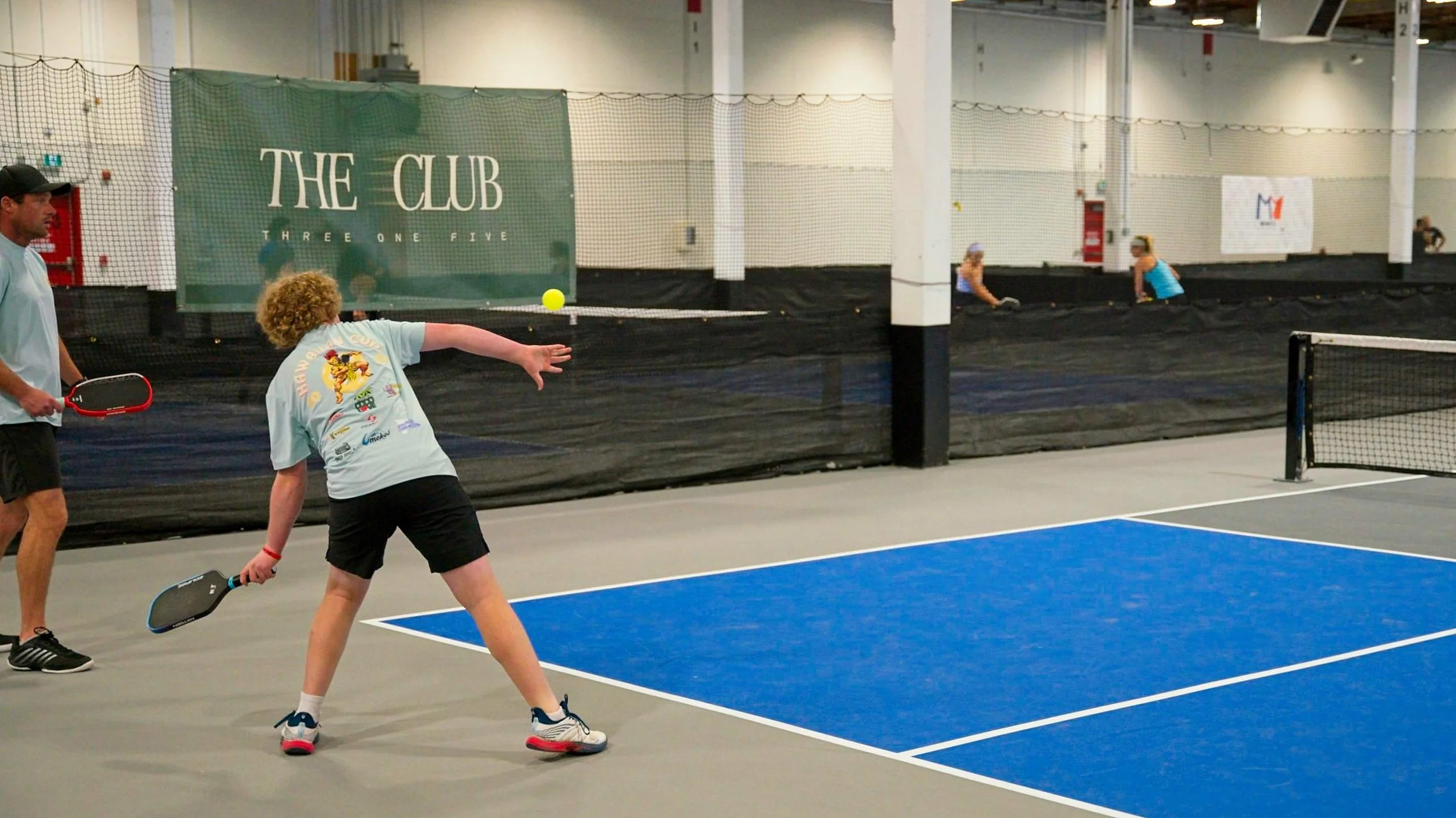 People playing pickleball on an indoor court with a blue playing surface, net, and adults with paddles engaged in the game.