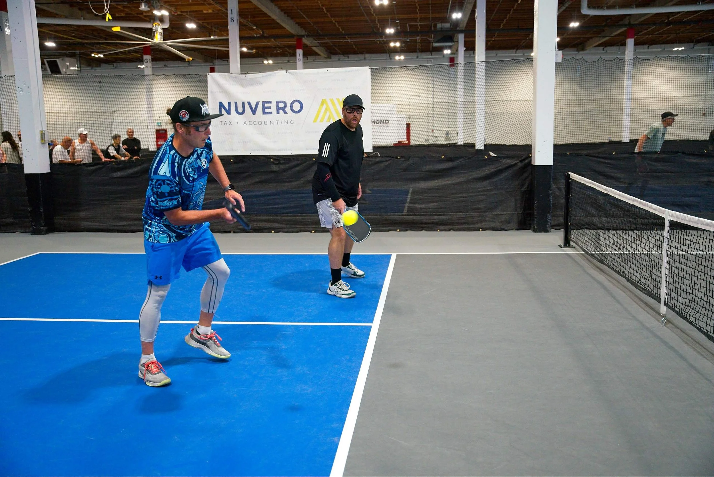 Two men are playing pickleball indoors on a court with blue and gray surfaces, with one man about to hit a yellow ball with a paddle while the other looks on. There are people in the background watching behind a net, and a large white banner with tex