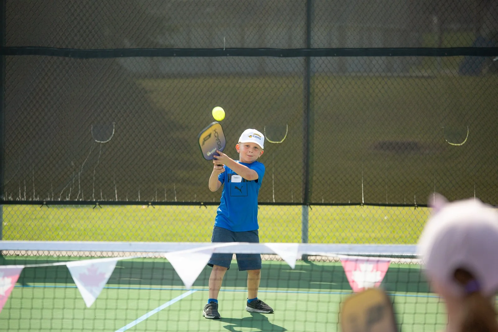 Young boy in blue shirt and white cap playing pickleball on an outdoor court, preparing to hit a yellow pickleball with a paddle.