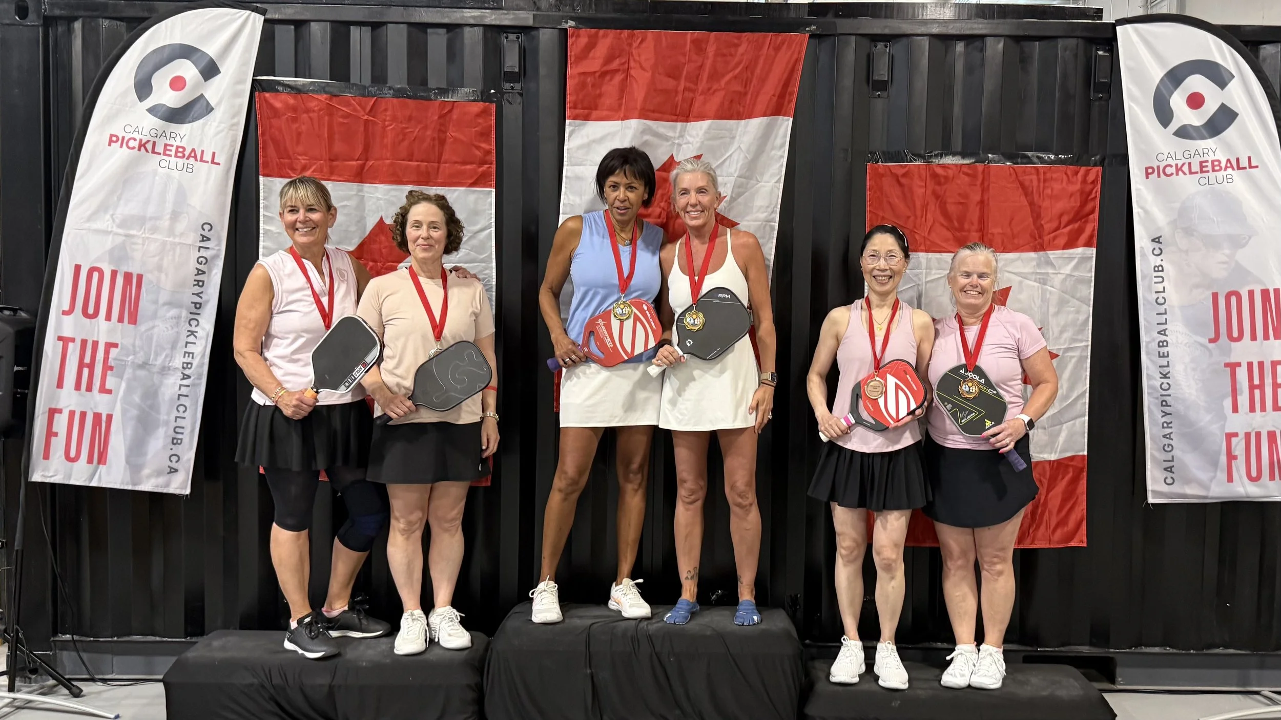 Group of women on a podium holding pickleball paddles, with Calgary Pickleball Club banners and flags in the background.