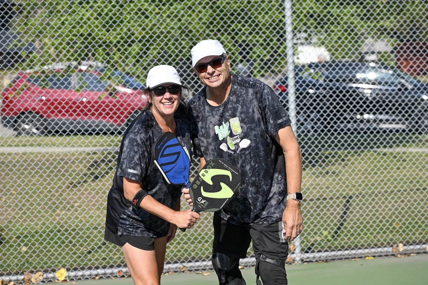Two people holding pickleball paddles on a pickleball court, smiling, wearing athletic clothing and hats, with a chain-link fence and parked cars in the background.