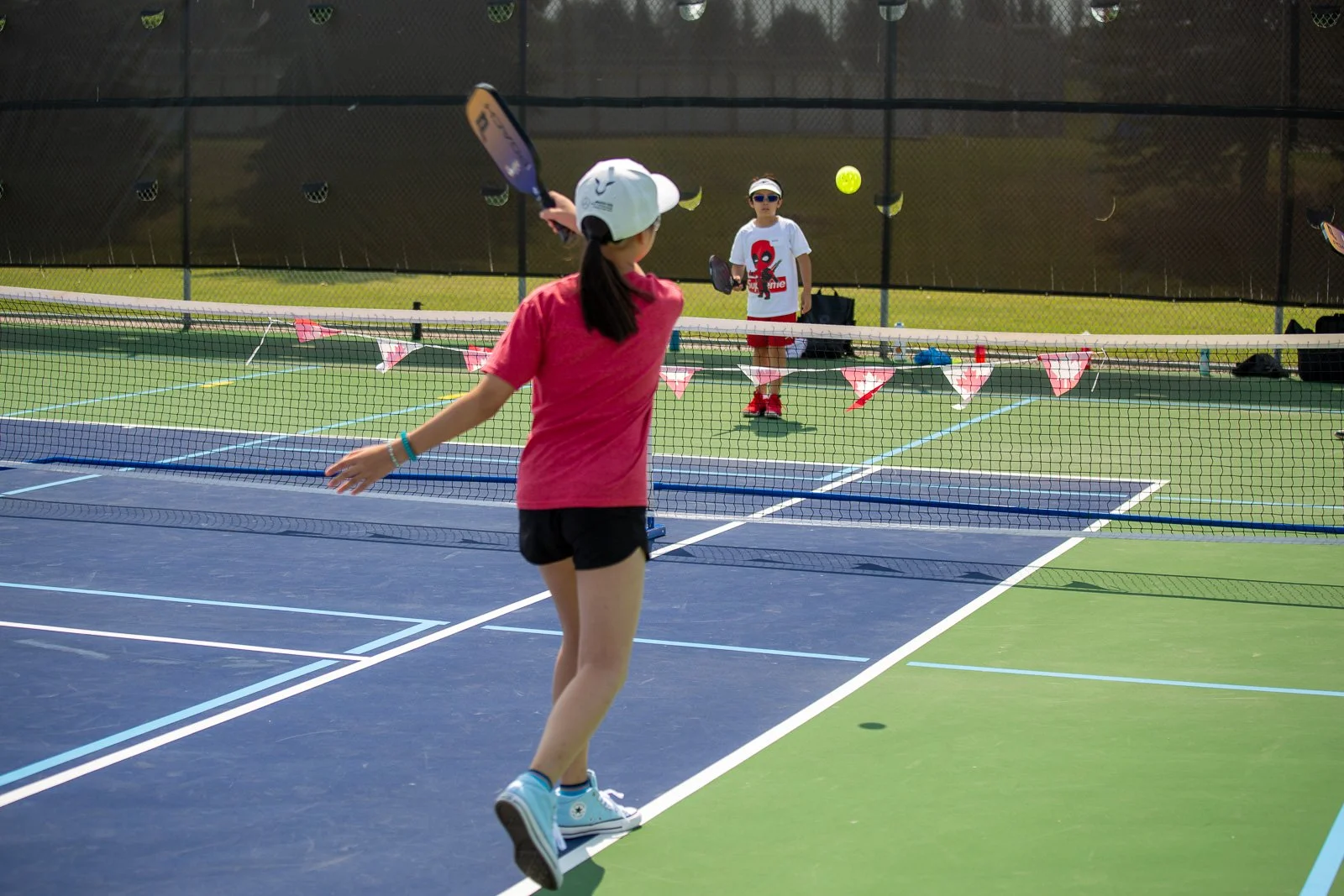 Children playing tennis on an outdoor court with a net and fencing, some holding tennis rackets and a tennis ball in the air.