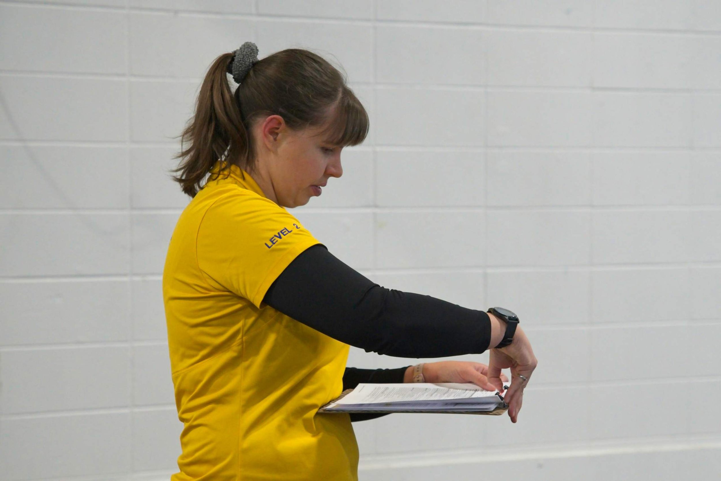 A woman wearing a yellow shirt and black long-sleeve shirt underneath, with a gray scrunchie in her brown hair tied back, is looking down and pointing at a clipboard with papers, standing against a white cinder block wall.