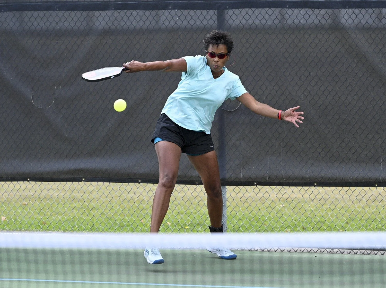 Woman playing tennis on a court, about to hit a yellow tennis ball with her racket, wearing sunglasses, a blue t-shirt, black shorts, and white tennis shoes.