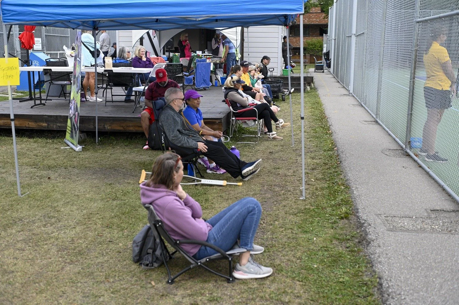 People sitting on chairs and benches outdoors near a tennis court, some wearing masks and a tent with volunteers behind them.