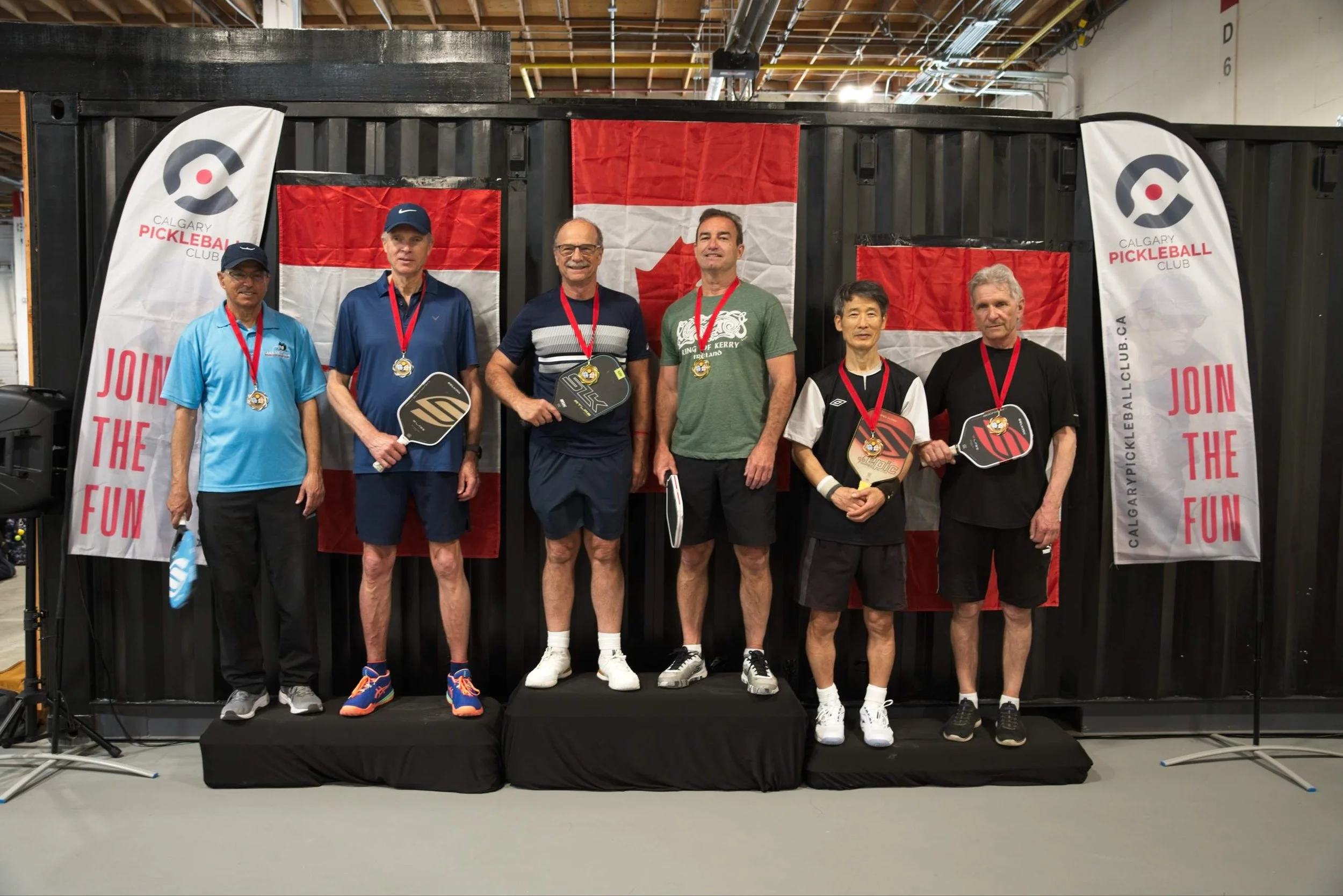 Group of six people standing on a small podium, holding pickleball paddles, wearing medals around their necks, in front of a black backdrop with red and white Canadian flags, and banners that say 'Join the Fun' and 'Calgary Pickleball Club'.