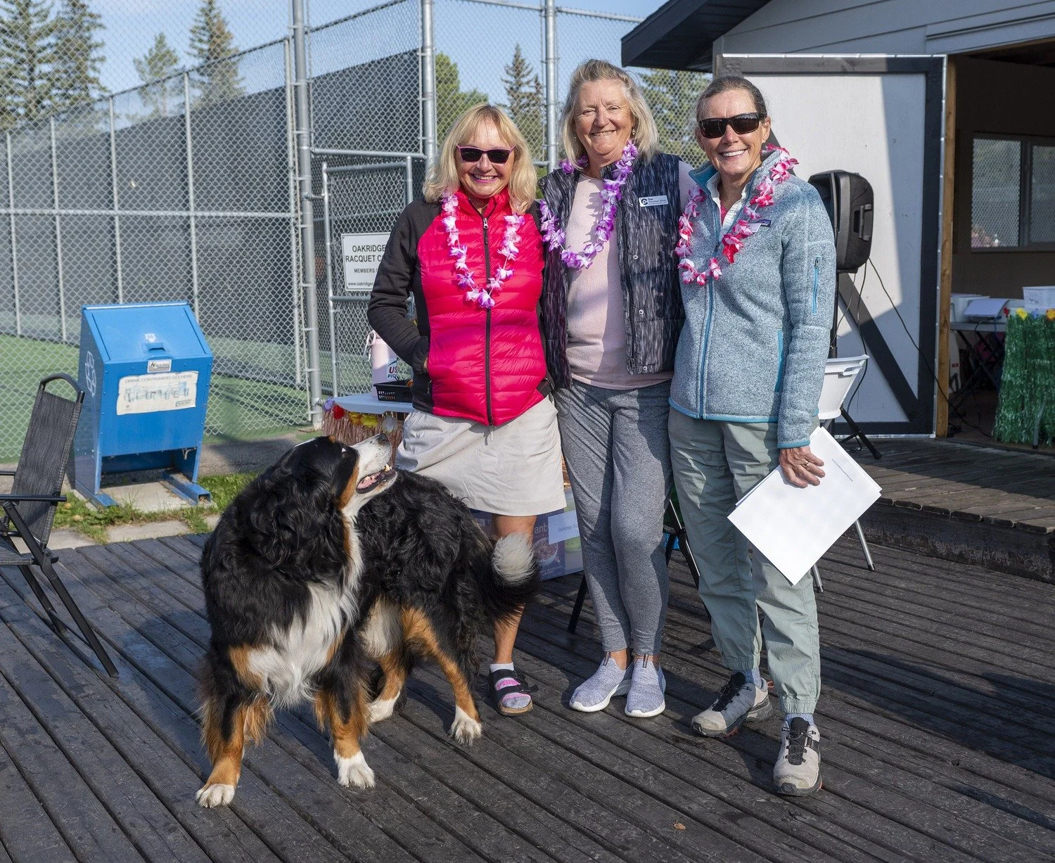 Three women standing on a wooden deck with a large Bernese mountain dog, all wearing pink and purple leis, at an outdoor tennis court.