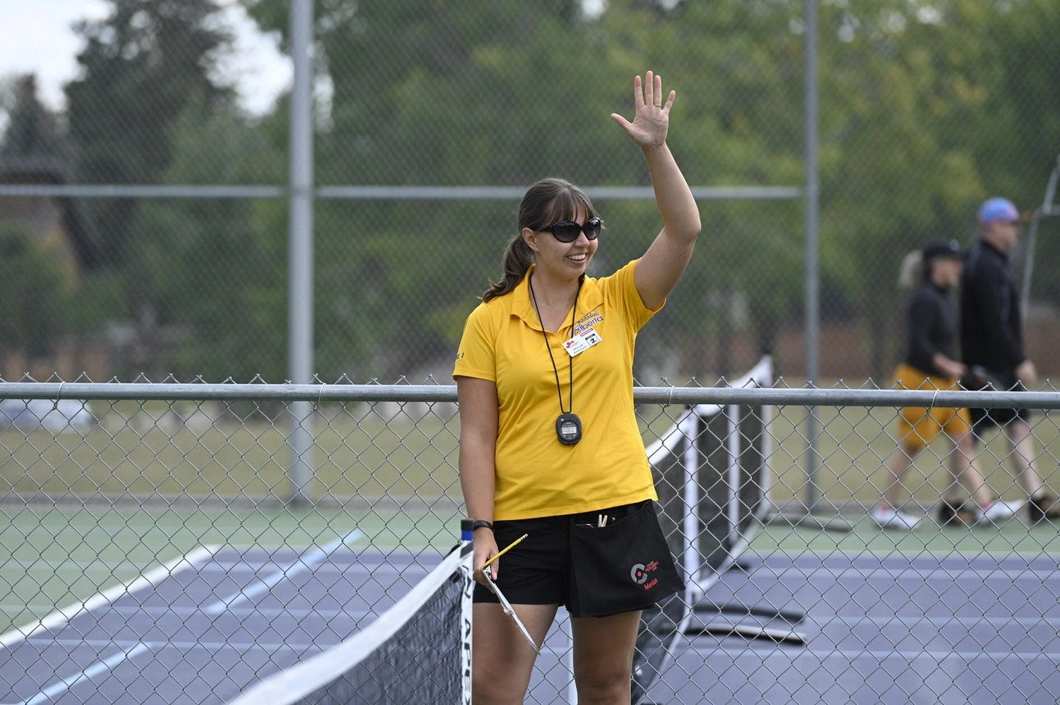 A woman in a yellow shirt and black shorts waving at a tennis court, wearing sunglasses and a whistle around her neck, with a few people in the background.