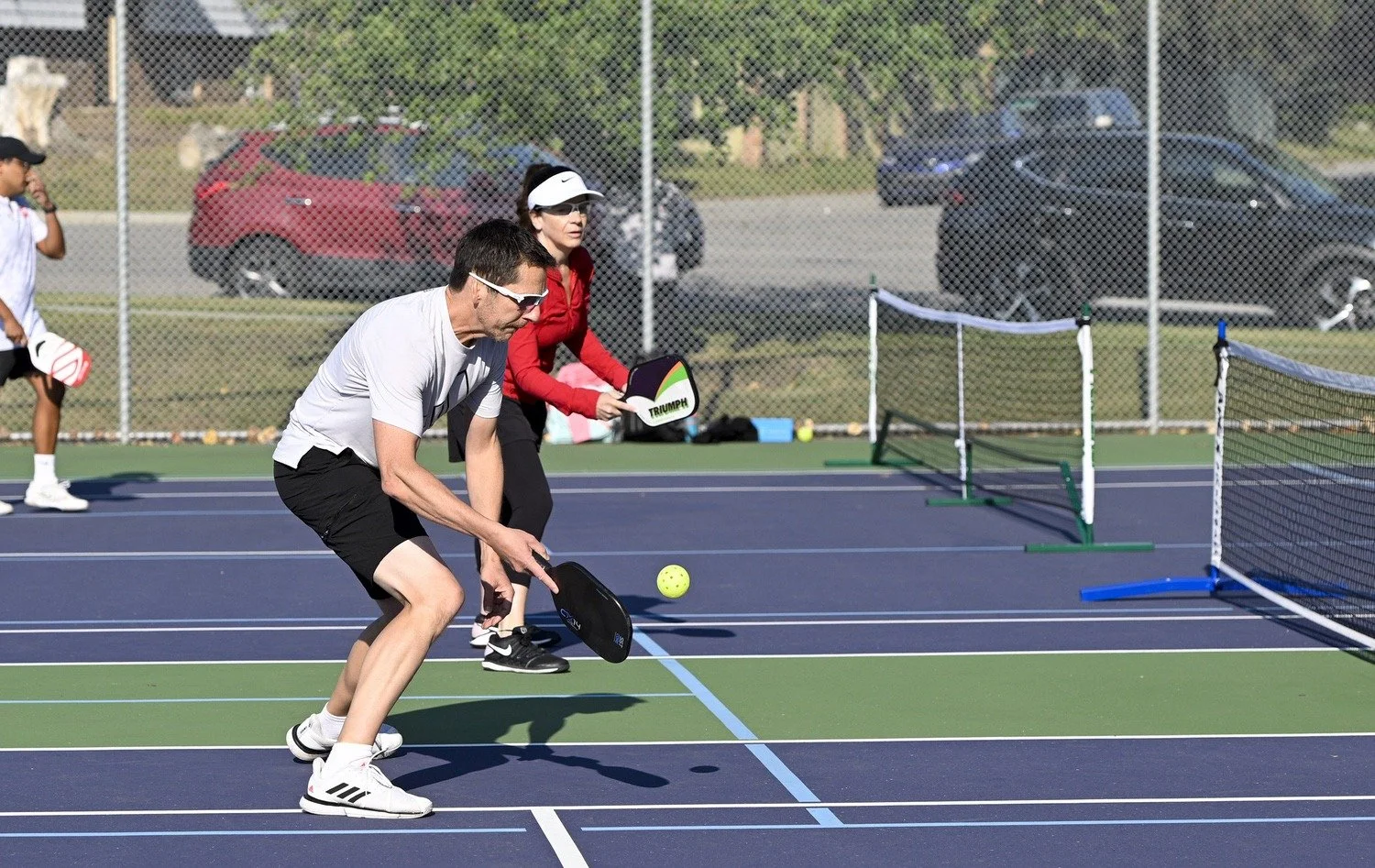 Two people play pickleball on an outdoor court with a blue surface and net, with a man in front hitting a ball with a paddle and a woman behind him holding a paddle, both wearing sunglasses and sportswear.