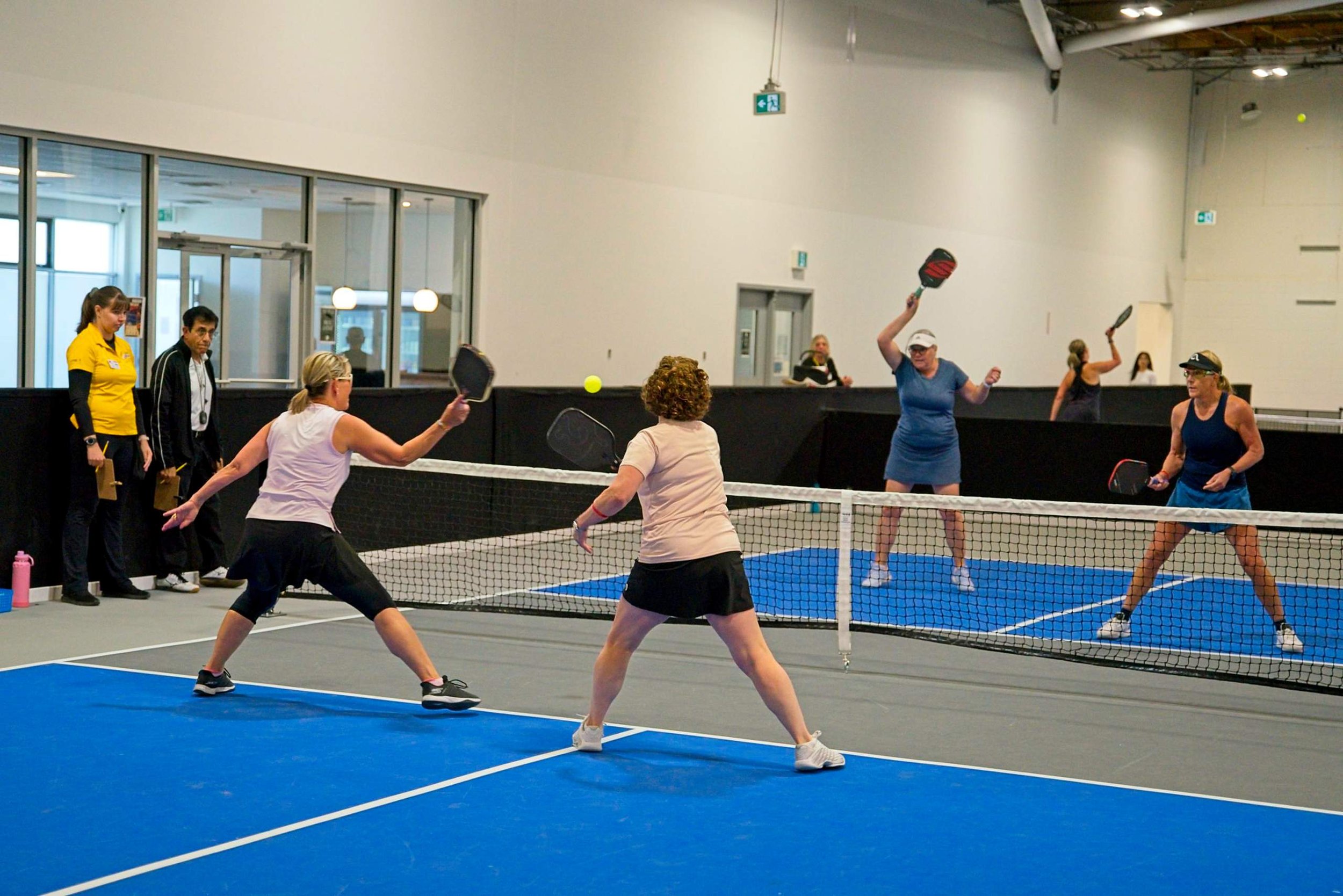 Women playing pickleball on an indoor court with a blue surface and a net, with spectators and staff observing.