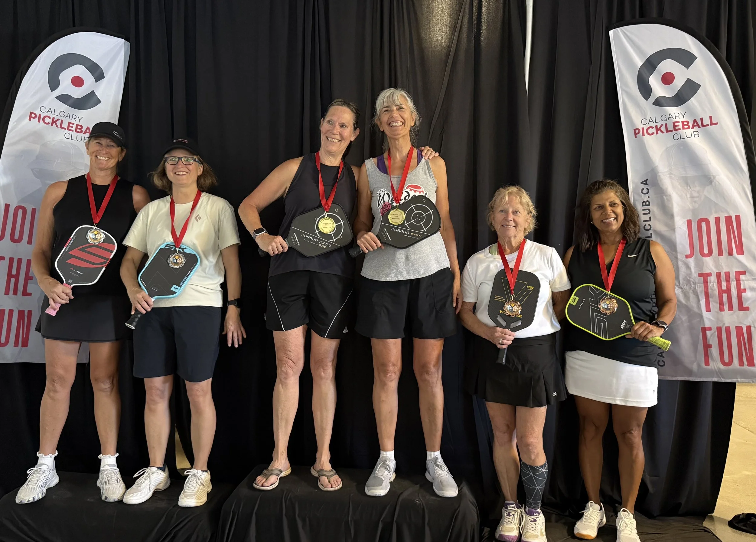 Group of seven women standing on a podium with medals, holding pickleball paddles, at the Calgary Pickleball Club event.