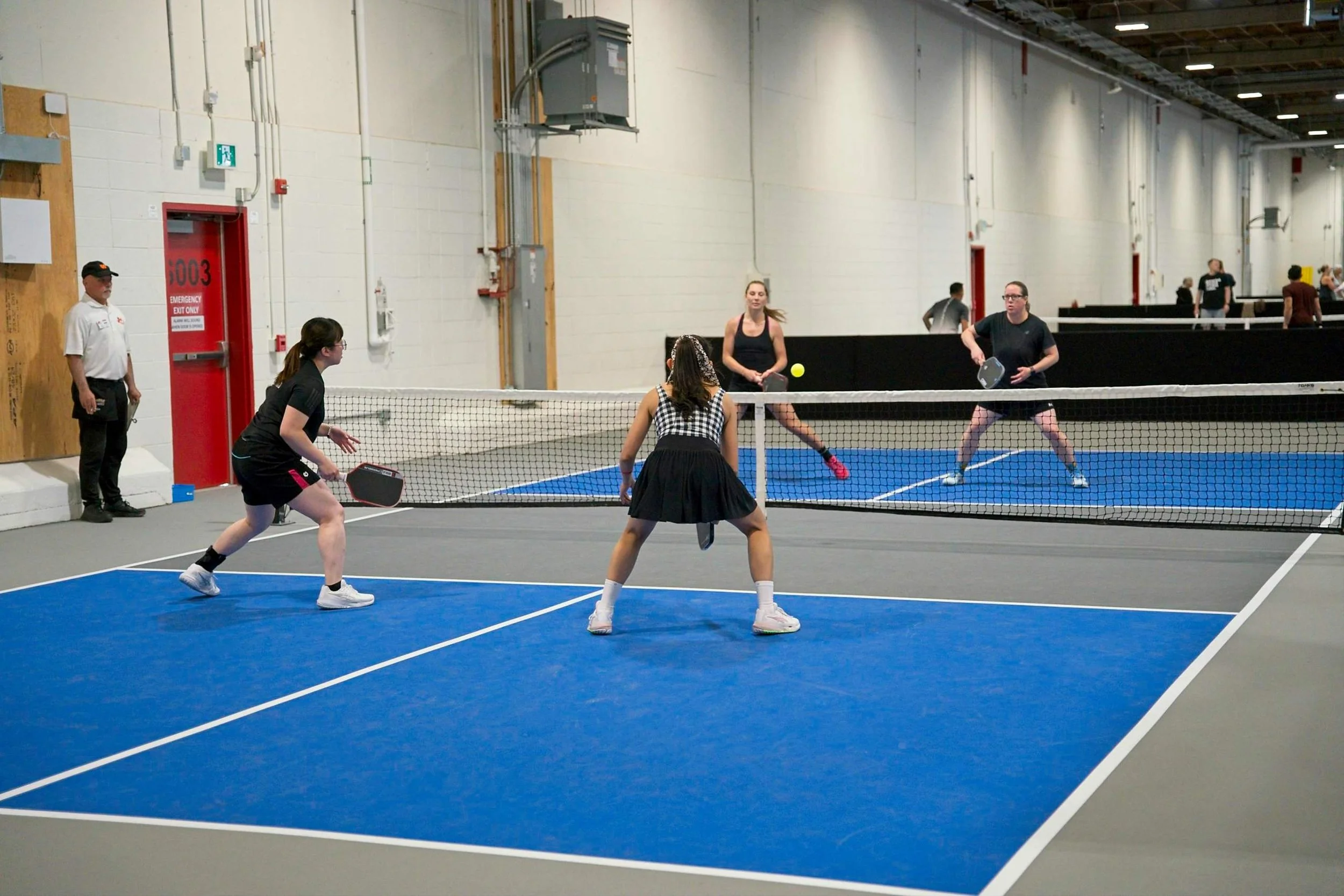 People playing pickleball inside a gymnasium with white walls, a black divider, and a blue court.
