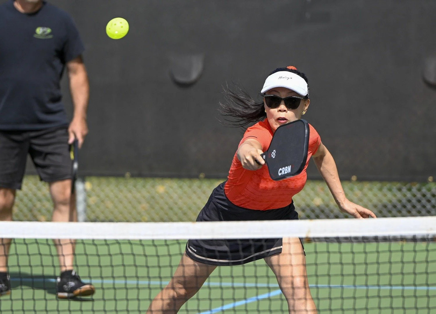 A woman playing pickleball, wearing sunglasses, a white visor, a red sports shirt, and black shorts, reaching to hit a bright yellow pickleball with a paddle. There is a man in the background on a pickleball court.