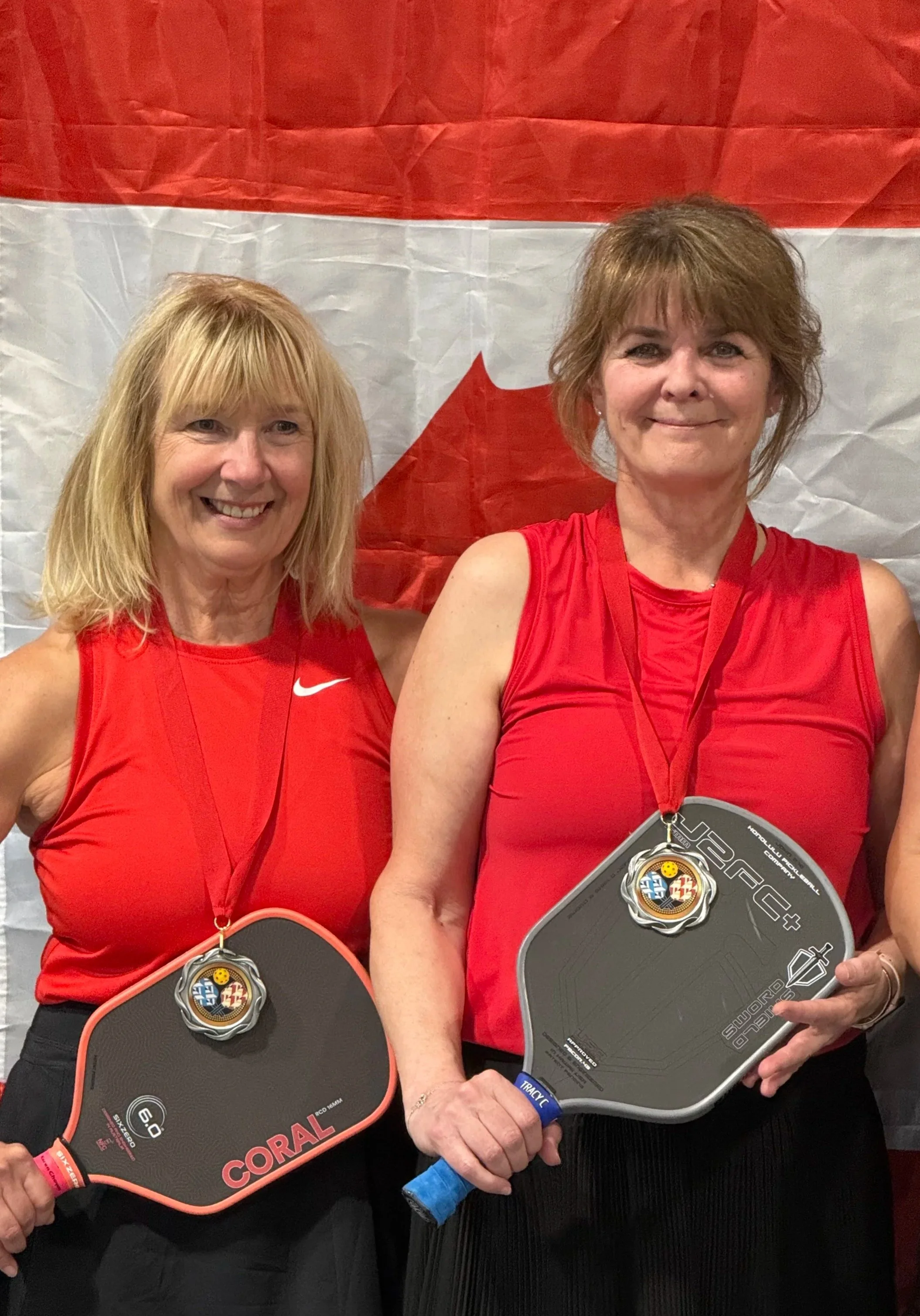 Two women in red sports shirts, holding paddle tennis rackets with medals, standing in front of a Canadian flag background.