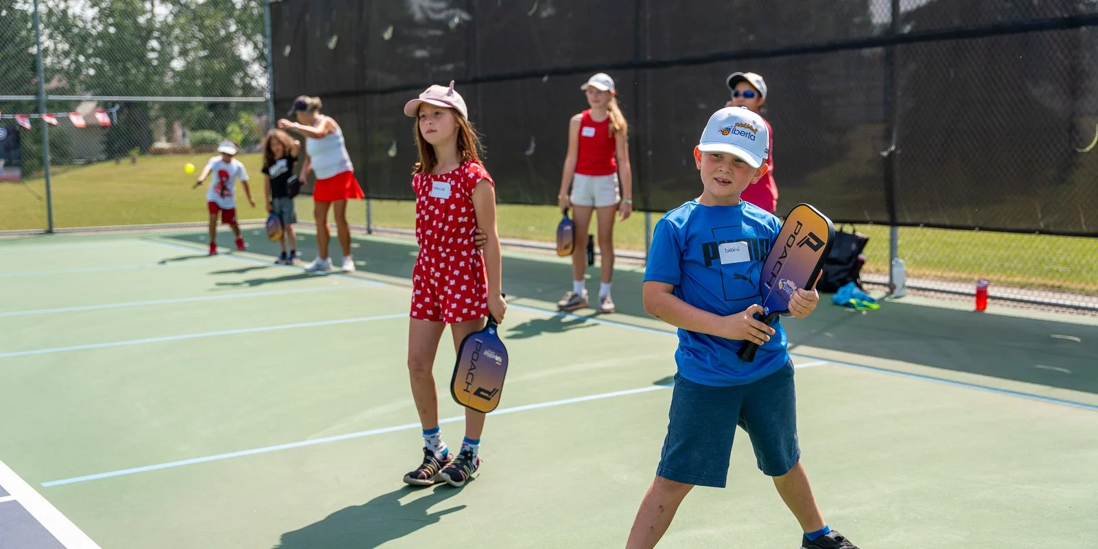 Children playing pickleball on an outdoor court, holding paddles, some wearing hats and sunglasses, with a chain-link fence in the background.