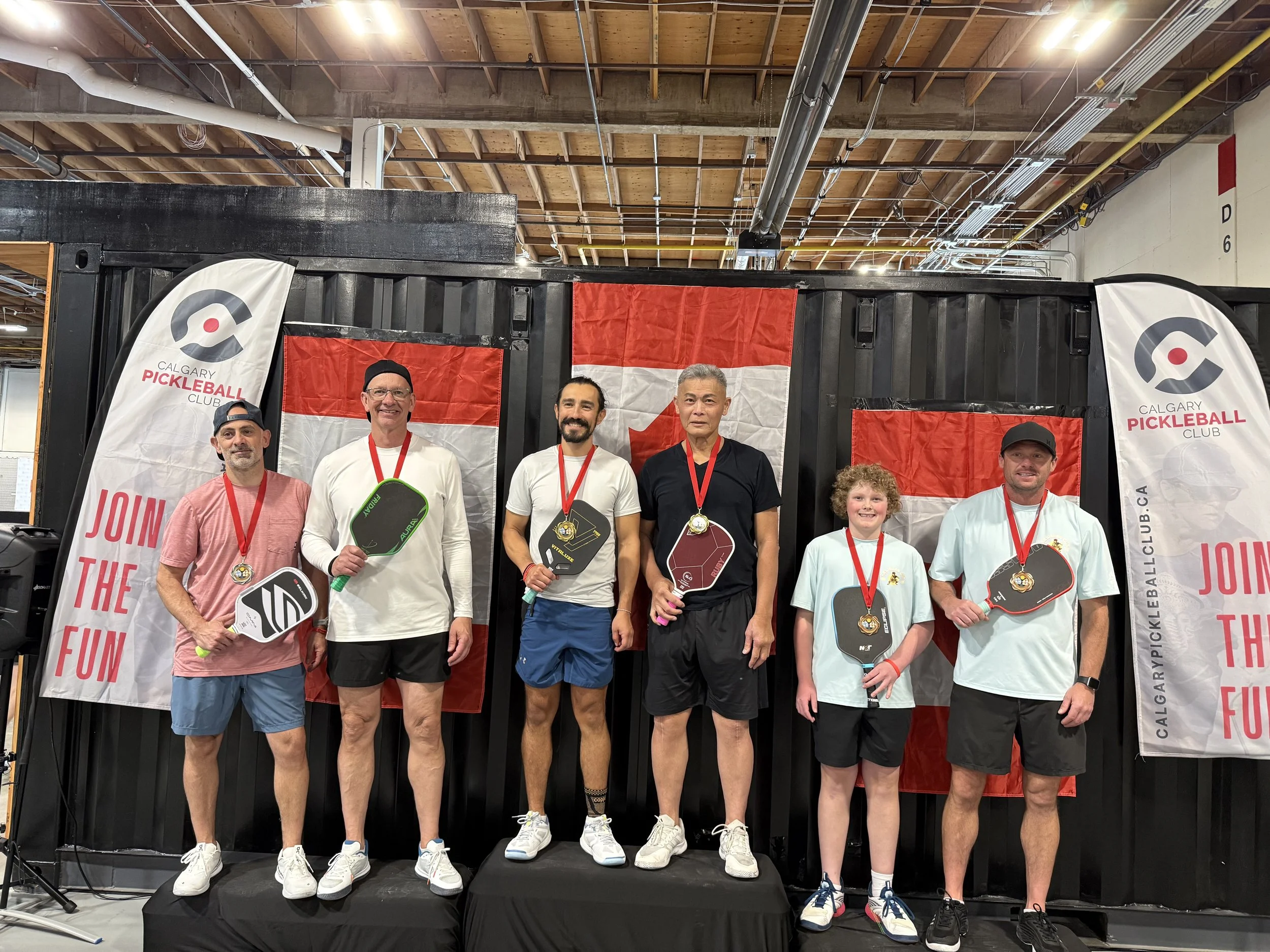 Group of five people standing on a winners' podium holding pickleball paddles at a Calgary Pickleball Club event. They are wearing medals and casual sportswear, with two flags of Calgary Pickleball Club and one Canadian flag in the background.